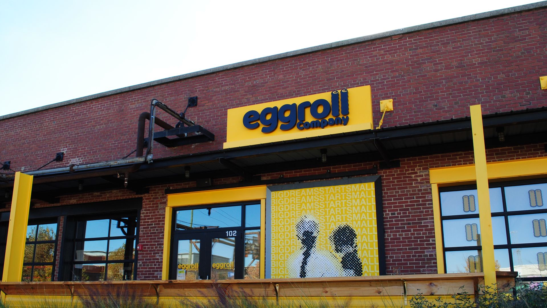 Brick building with a bright yellow sign reading "eggroll company" above black-framed glass door and windows. Yellow panel with text "BIG DREAMS START SMALL" and silhouettes of two people.