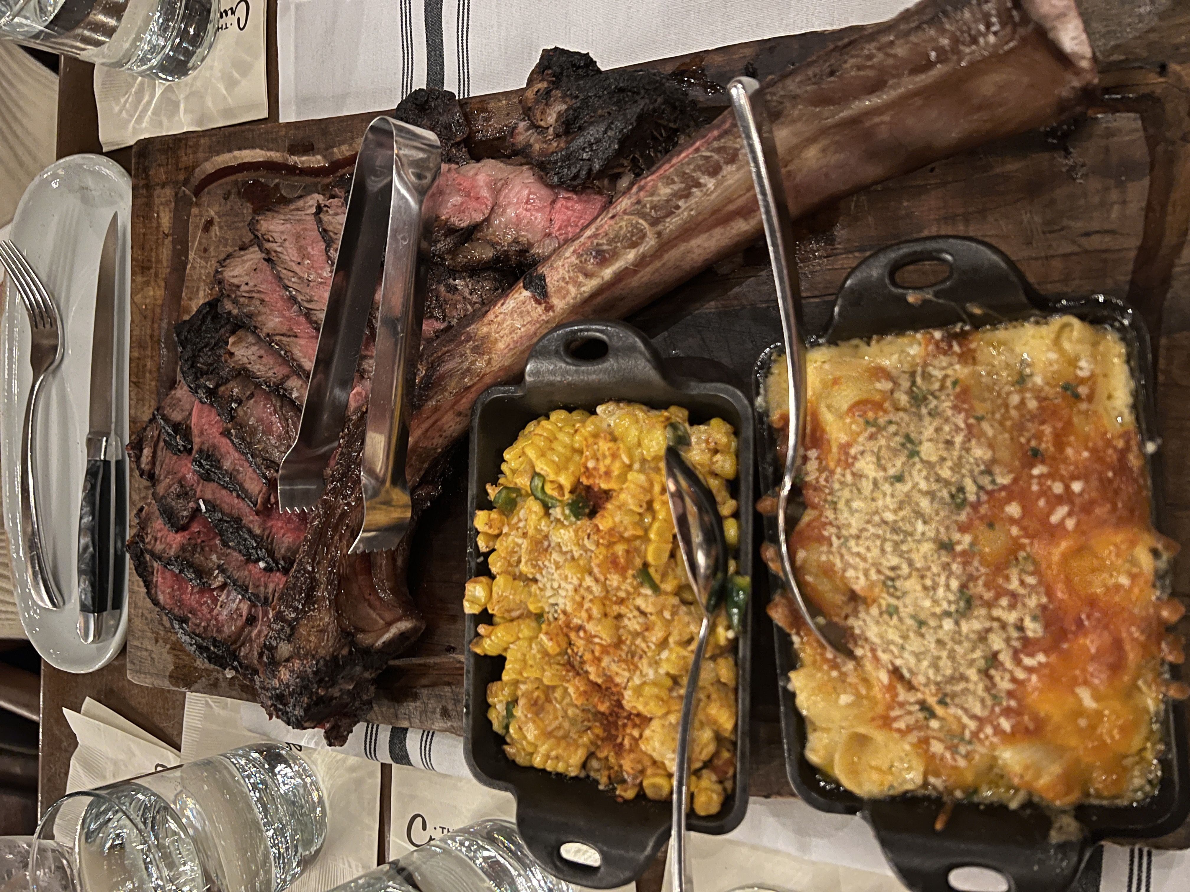 A large and juicy tomahawk steak is served on a wooden cutting board, with several slices cut and fanned out. Next to the steak, there are two small cast iron skillets. One contains a cheesy corn dish, and the other is filled with mac and cheese topped with breadcrumbs. 