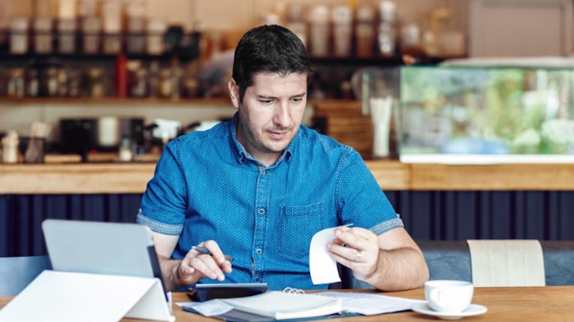 A man sitting at a table looking at a receipt and using a calculator. He also has a tablet, a cup, and various papers in front of him.