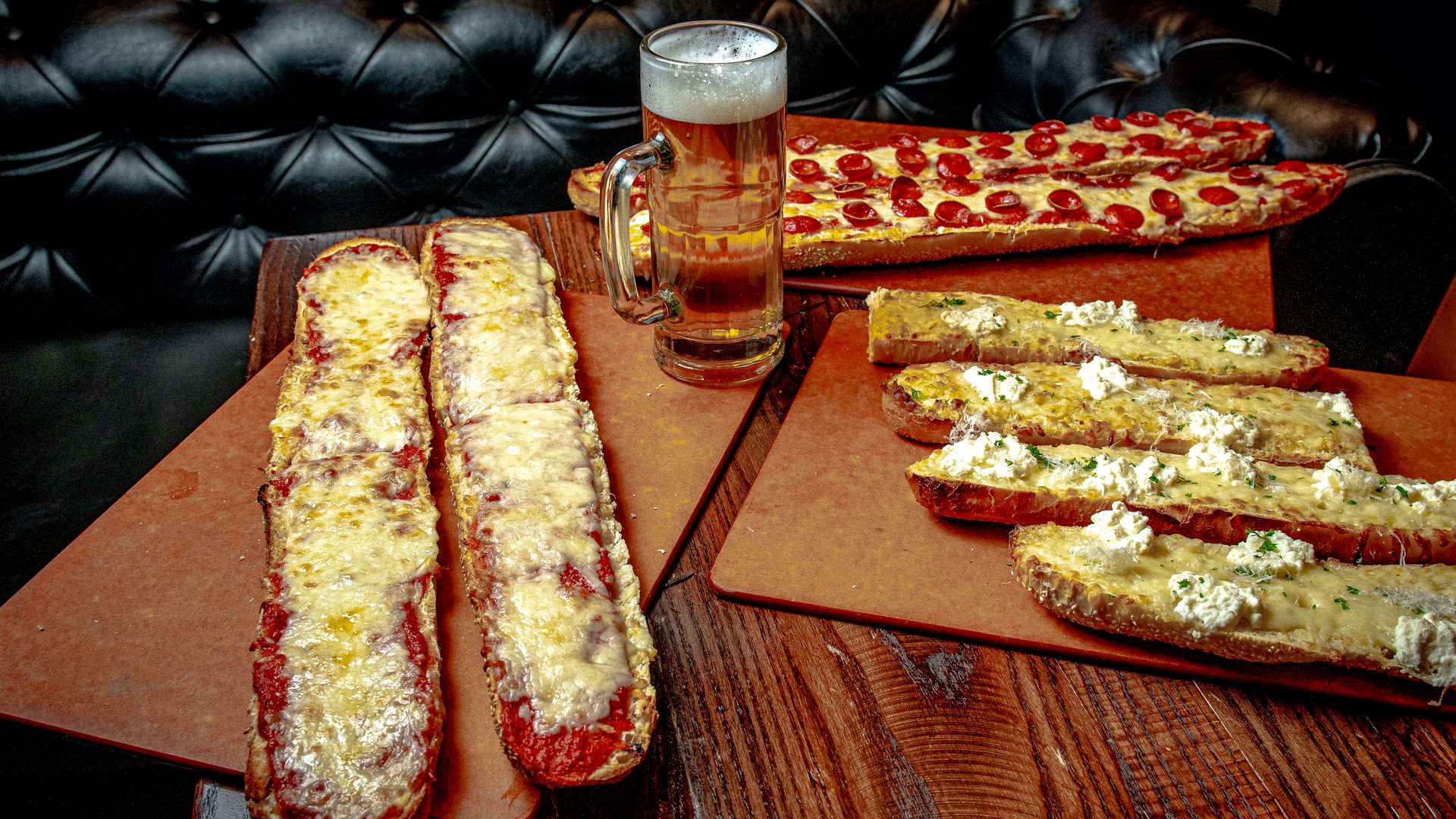 French bread pizzas, including cheese and pepperoni, on a table with a tall mug of beer and black banquette in the background