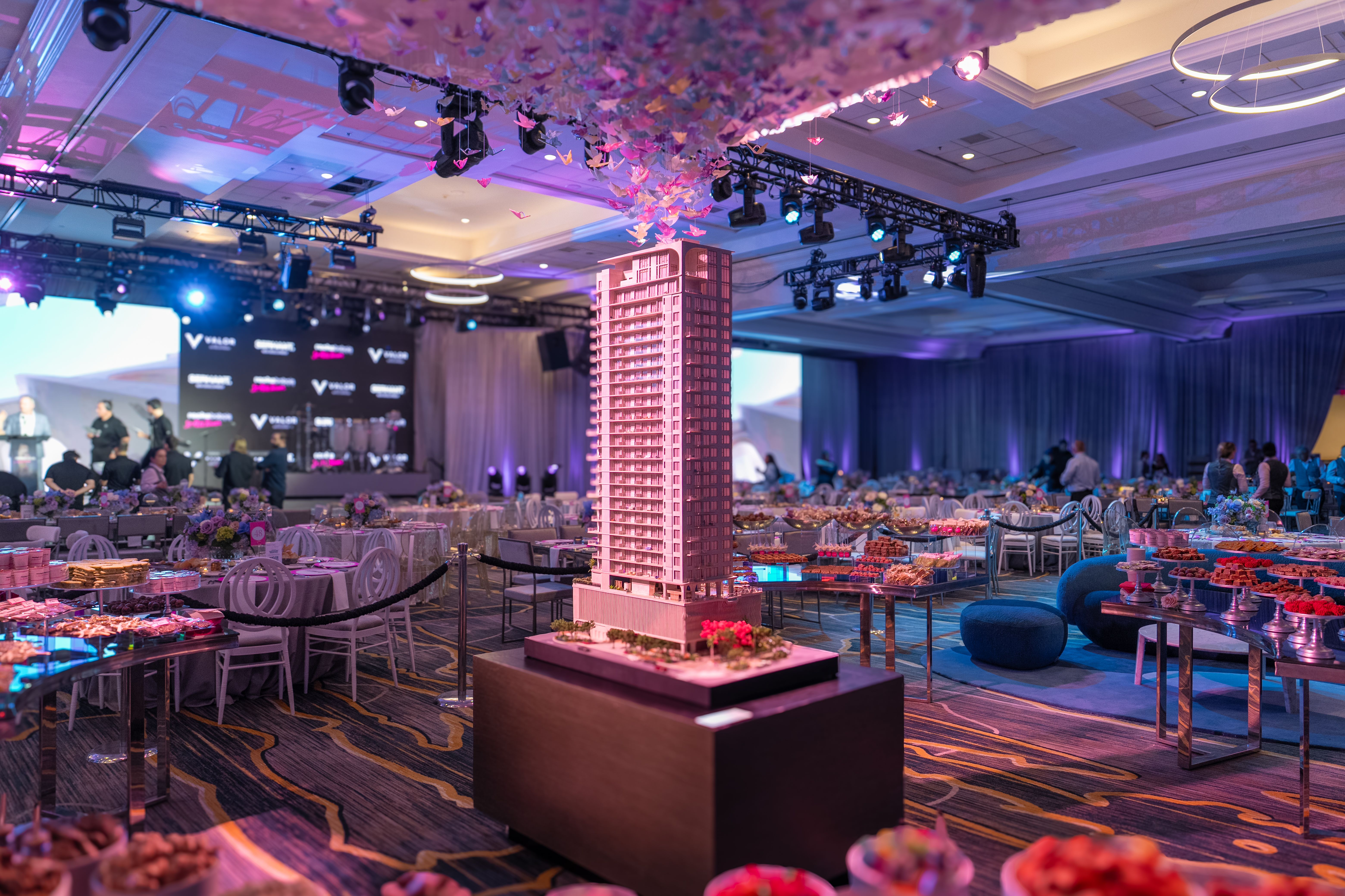Indoor event with a tall architectural building model in the center, surrounded by tables with white chairs and floral centerpieces, under pink and purple ambient lighting.