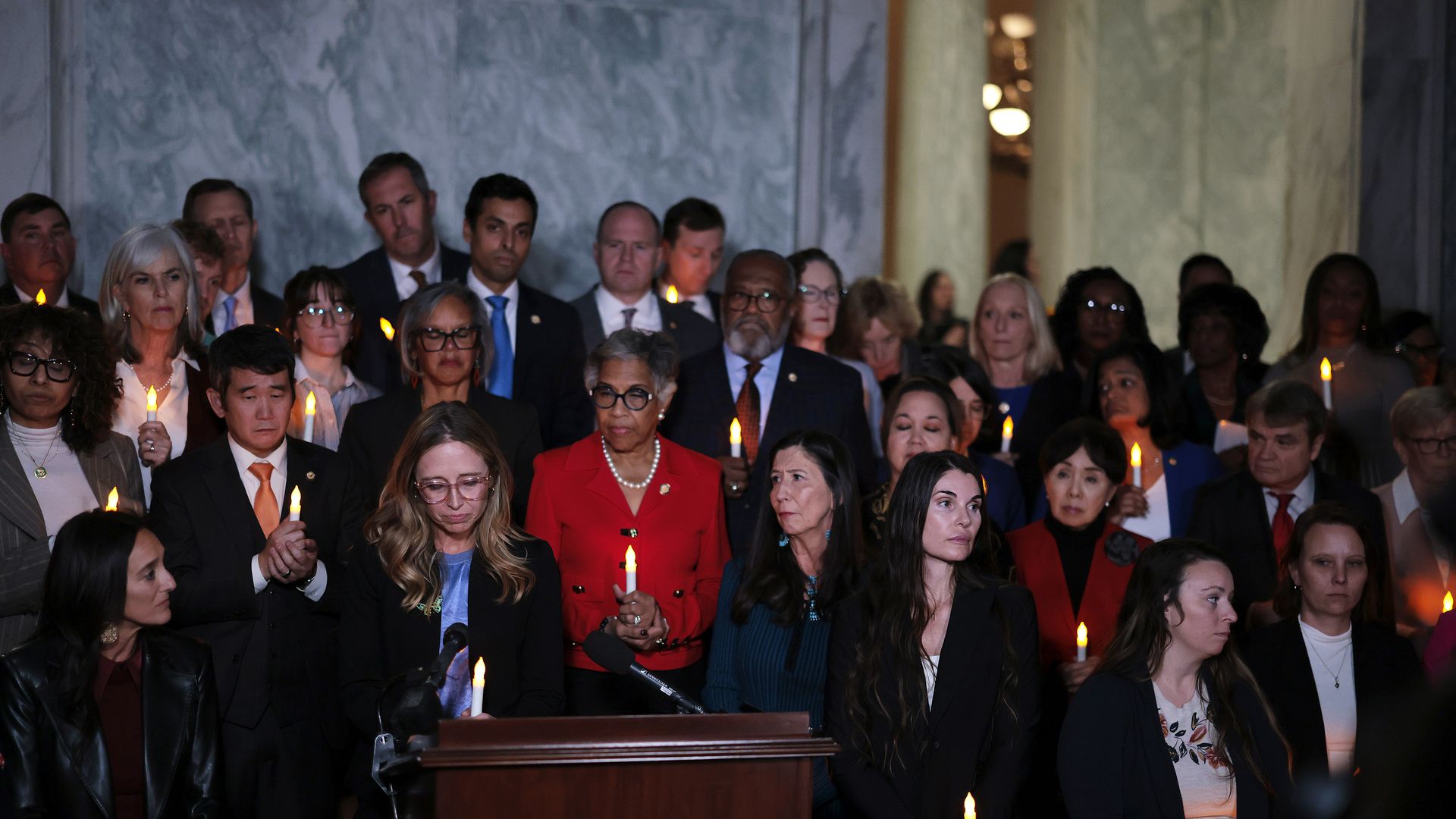 A group of men and women hold candles in their hands, while some look down solemnly