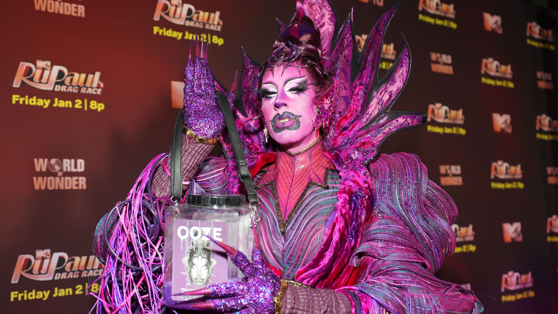 Person in elaborate purple and pink costume with spiked headpiece and glittery gloves holds a clear container labeled OOTE, posing at "RuPaul's Drag Race" event backdrop.
