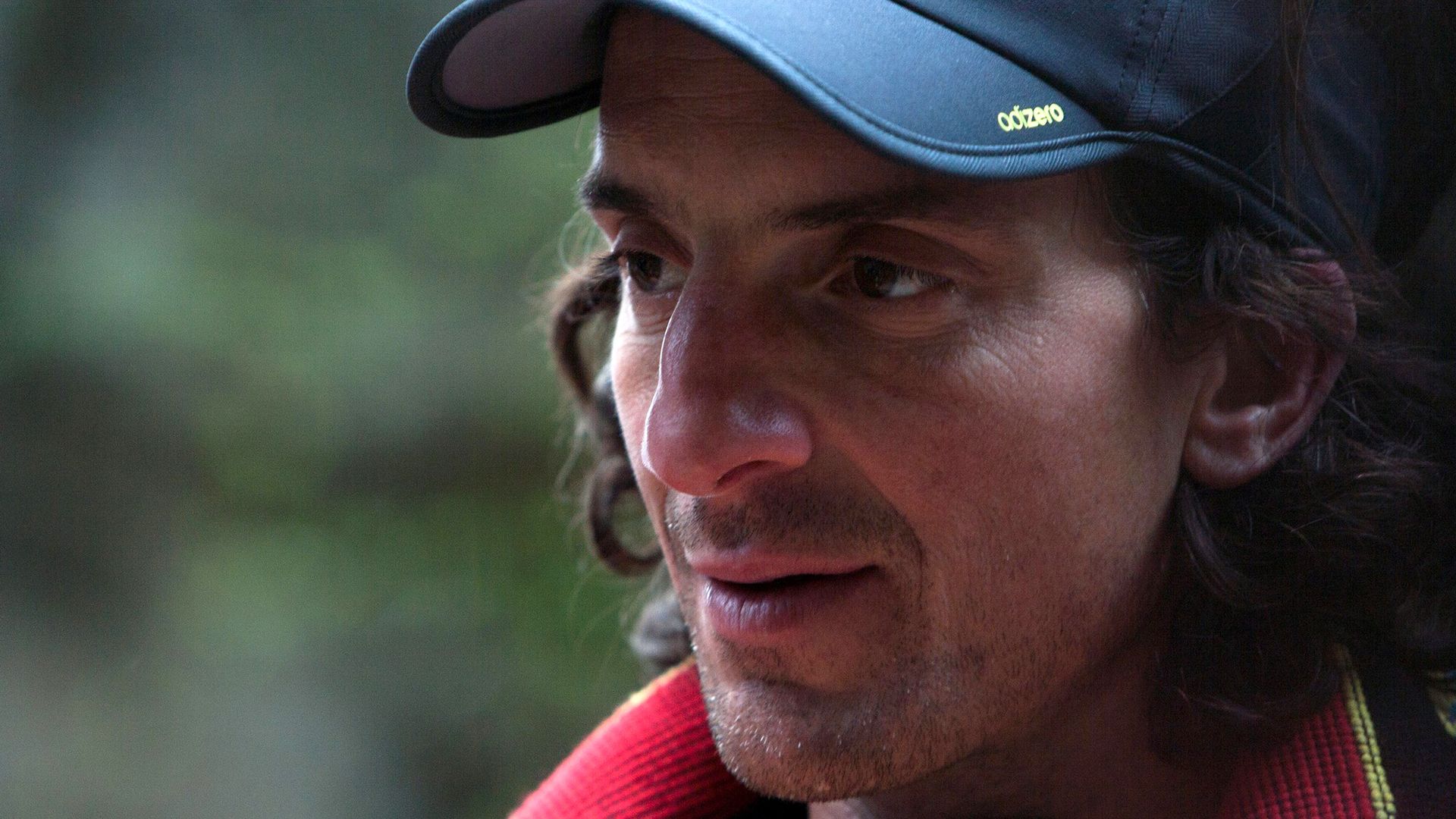 Close-up of a man with long dark hair and light stubble, wearing a dark blue cap and red outerwear, looking to the left, with a blurred green outdoor background.