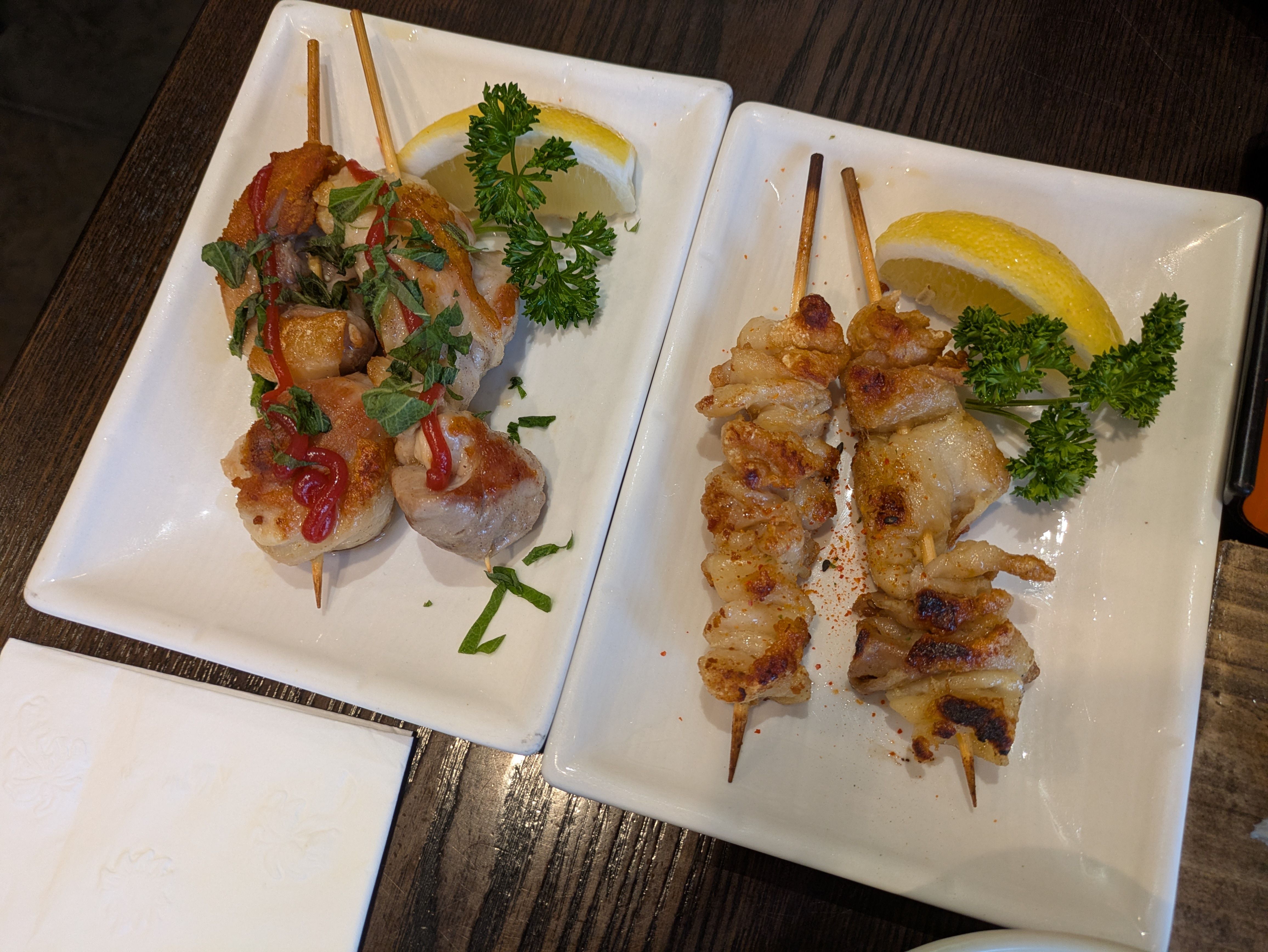 Two white plates on a dark wooden table hold grilled skewers. Left: herb-topped meatball skewers with red sauce and a lemon. Right: chicken skewers with onions, lemon, and parsley.