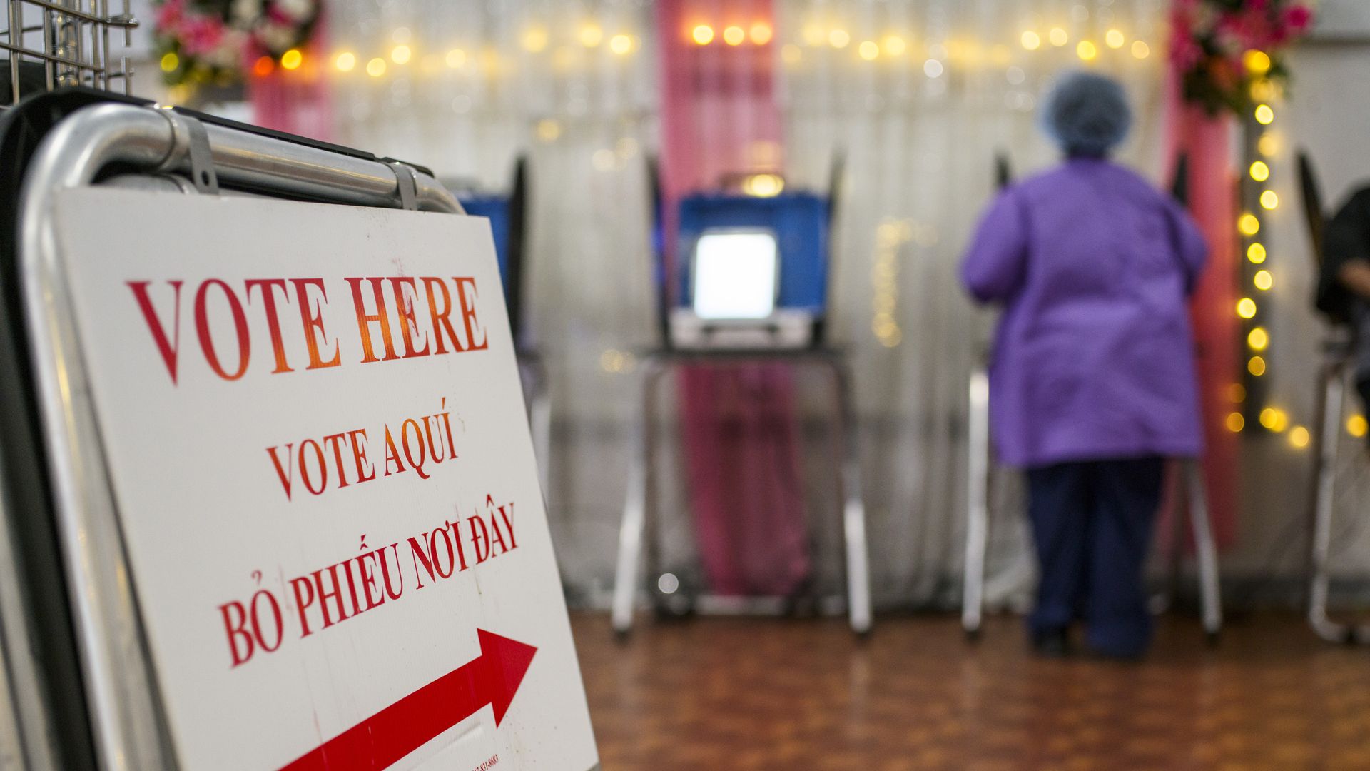 A multilingual voting sign with red text and an arrow directing to voting booths, and a person in a purple coat standing at one booth in a decorated room with wooden flooring.