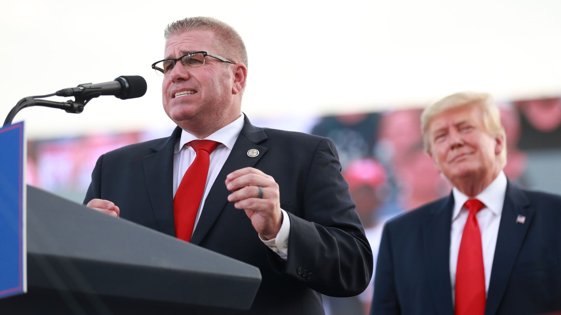 Man in a dark suit and red tie speaking at a microphone, with another man in a similar outfit standing blurred in the background, both at an outdoor event.
