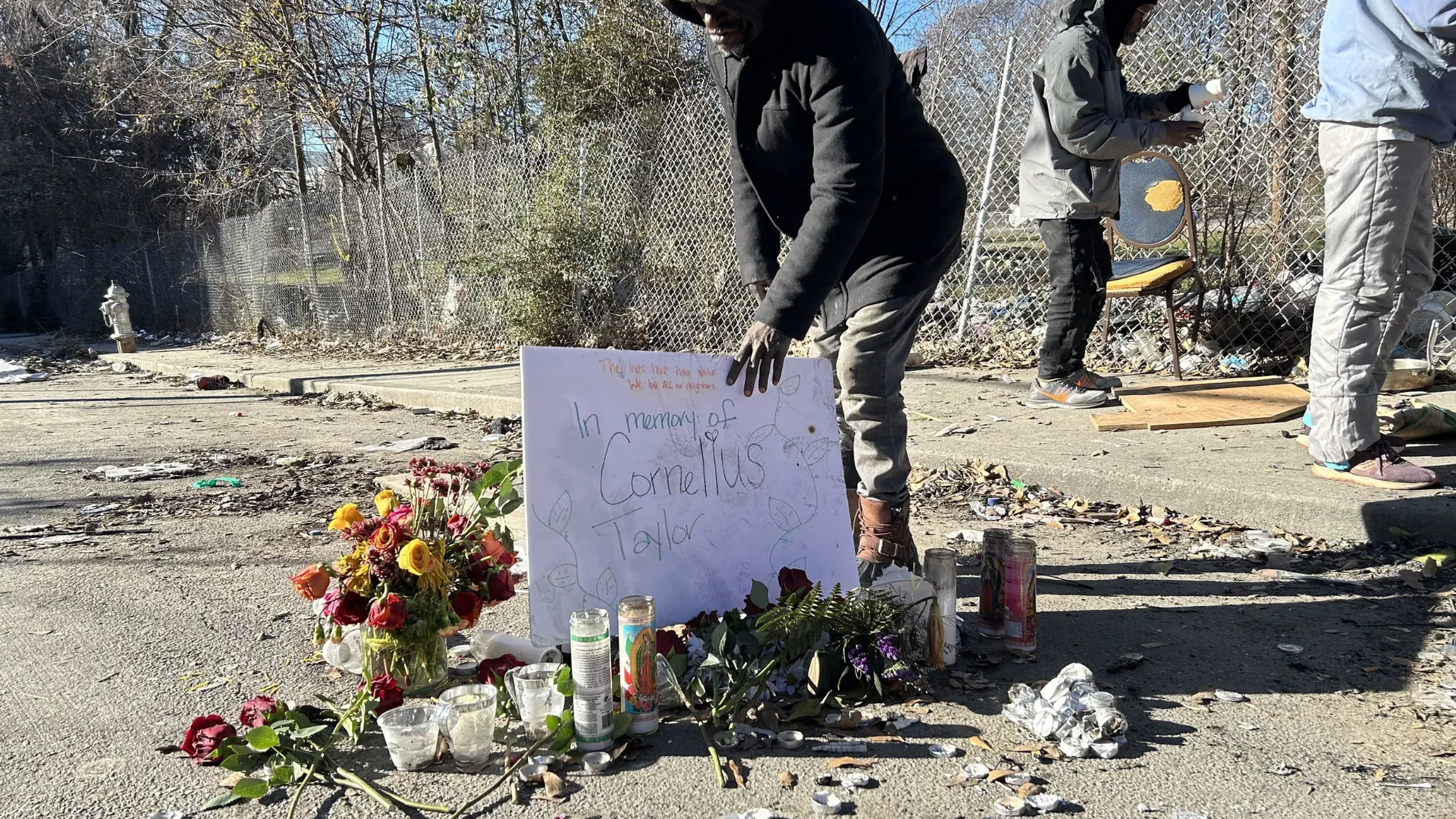 A person placing a memorial sign that reads "In memory of Cornelius Taylor" at a roadside tribute with flowers and candles, next to a chain-link fence on a sunny day.