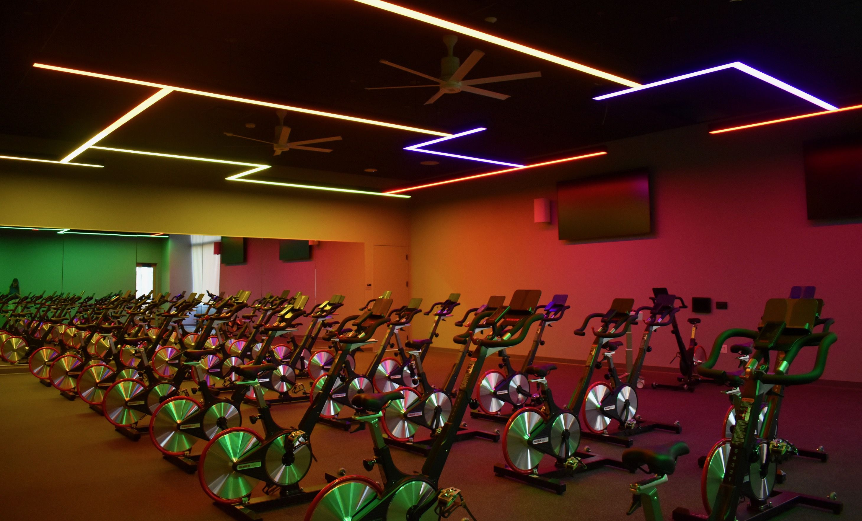Indoor spin class room with rows of stationary bikes under colorful neon ceiling lights in green, yellow, orange, purple, and red hues, and wall mirrors reflecting the bikes.