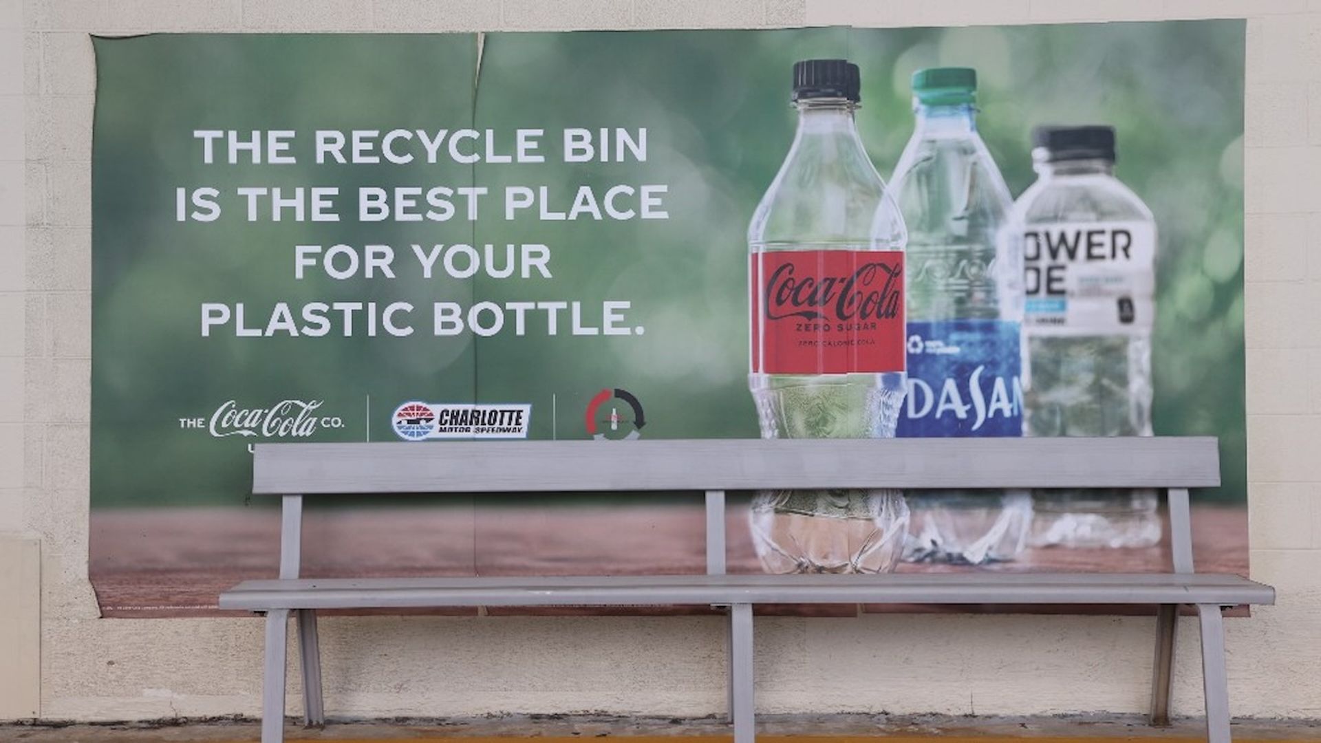 A bench sits in front of a recycling awareness poster showing plastic bottles and the message, "The recycle bin is the best place for your plastic bottle."