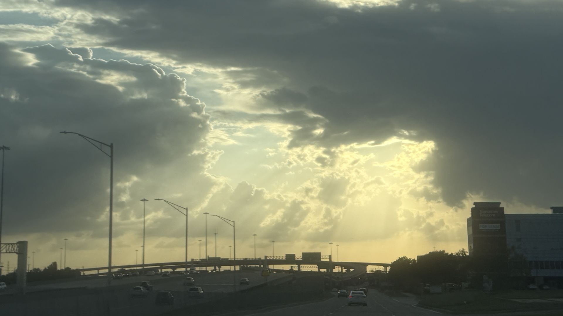 A highway scene at sunset with dark storm clouds and bright sunbeams streaming through. Streetlights line the road as cars travel; a large building sits to the right.