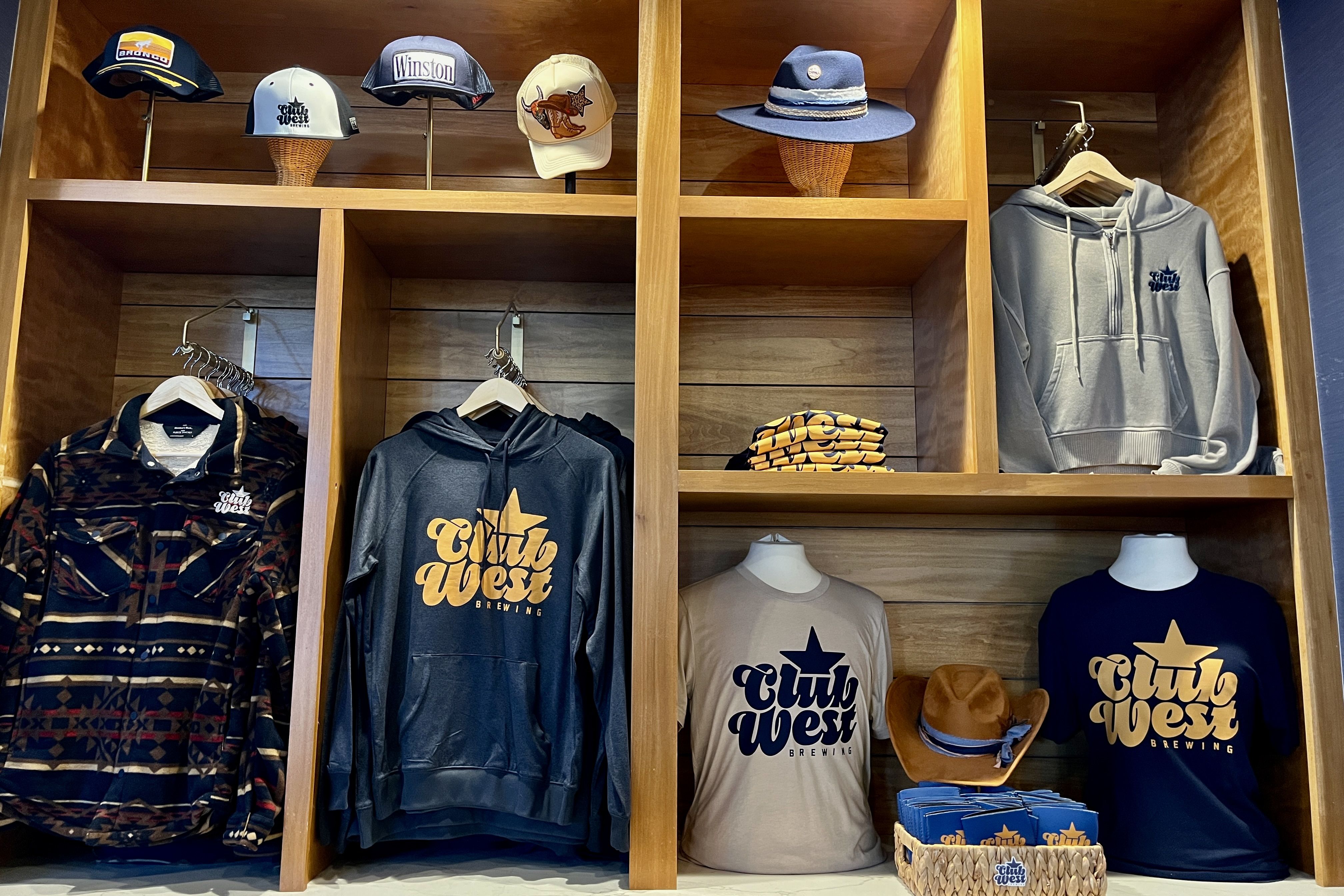 Wooden cubby display of City West Brewing apparel: top hats and caps; middle plaid jacket and gray hoodies; bottom beige tee with star, navy tee, and a brown cowboy hat.