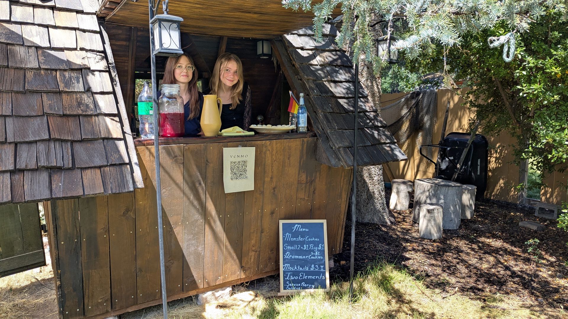 Two girls sell cold drinks and cookies from a small wooden shack.