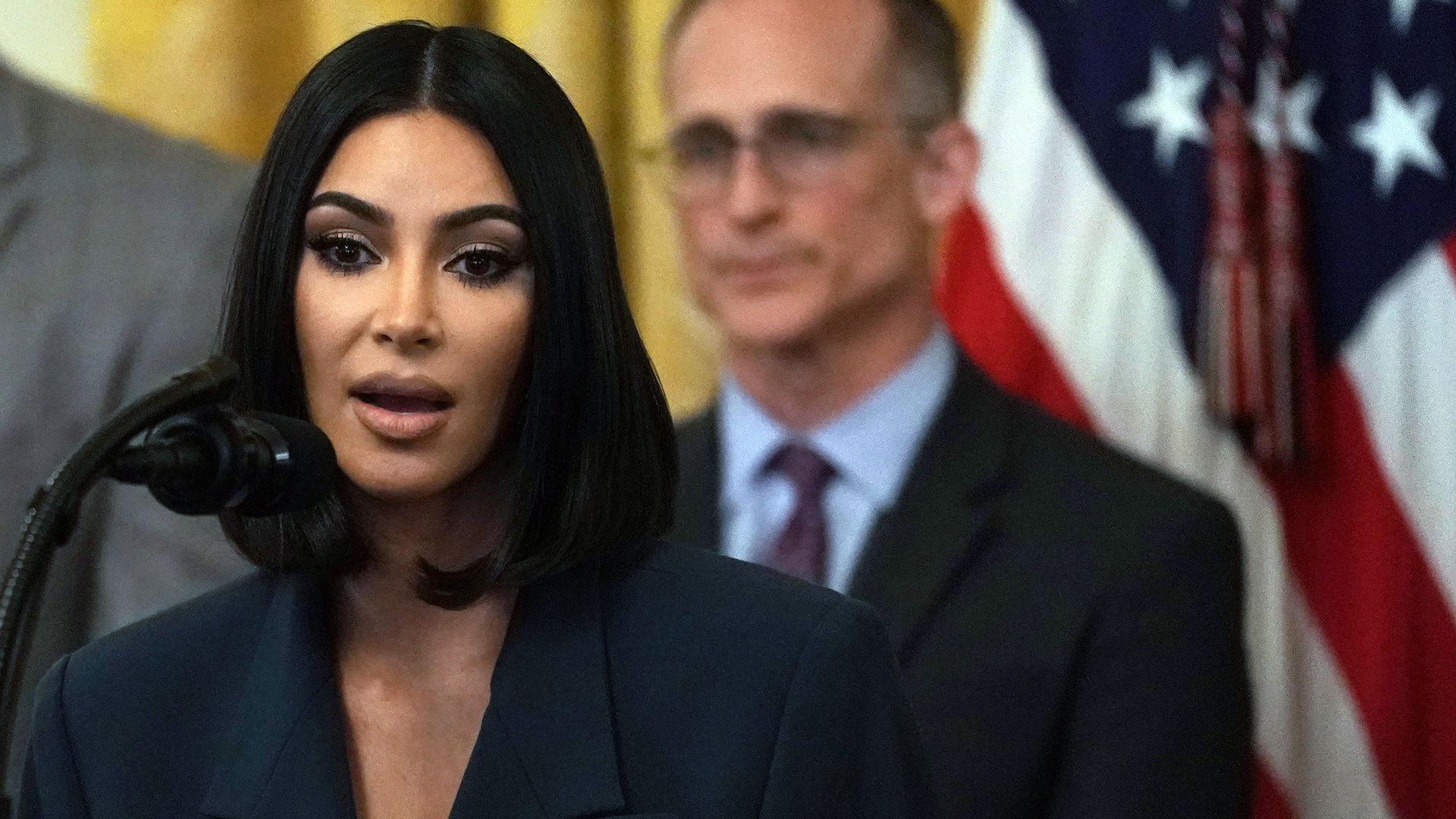 Kim Kardashian speaks at a White House event with an American flag in the background.