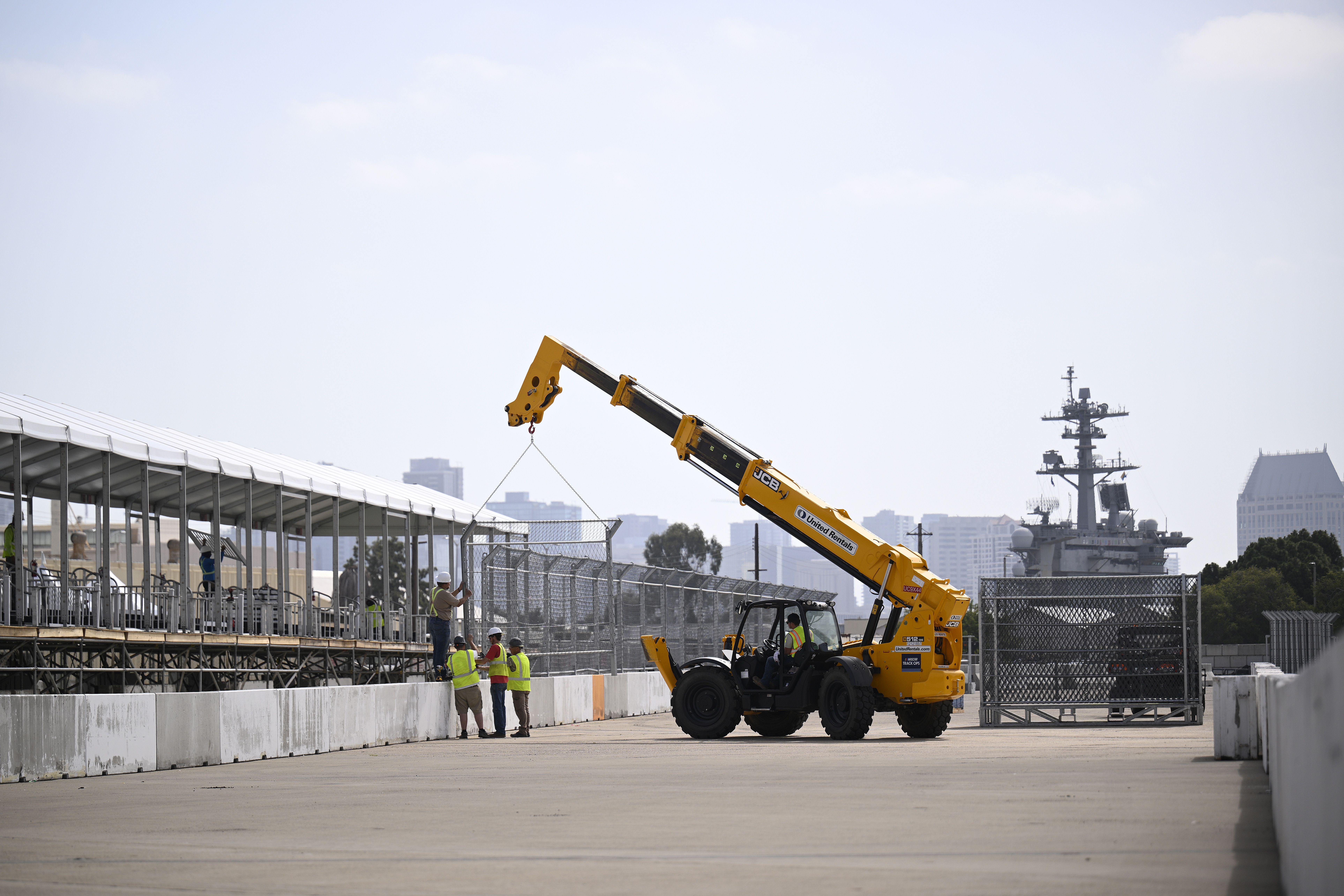 A construction site with a yellow crane arm lifting a load, workers in hi-visibility vests and hard hats, scaffolding along spectator stands, and an aircraft carrier and city skyline in the background.
