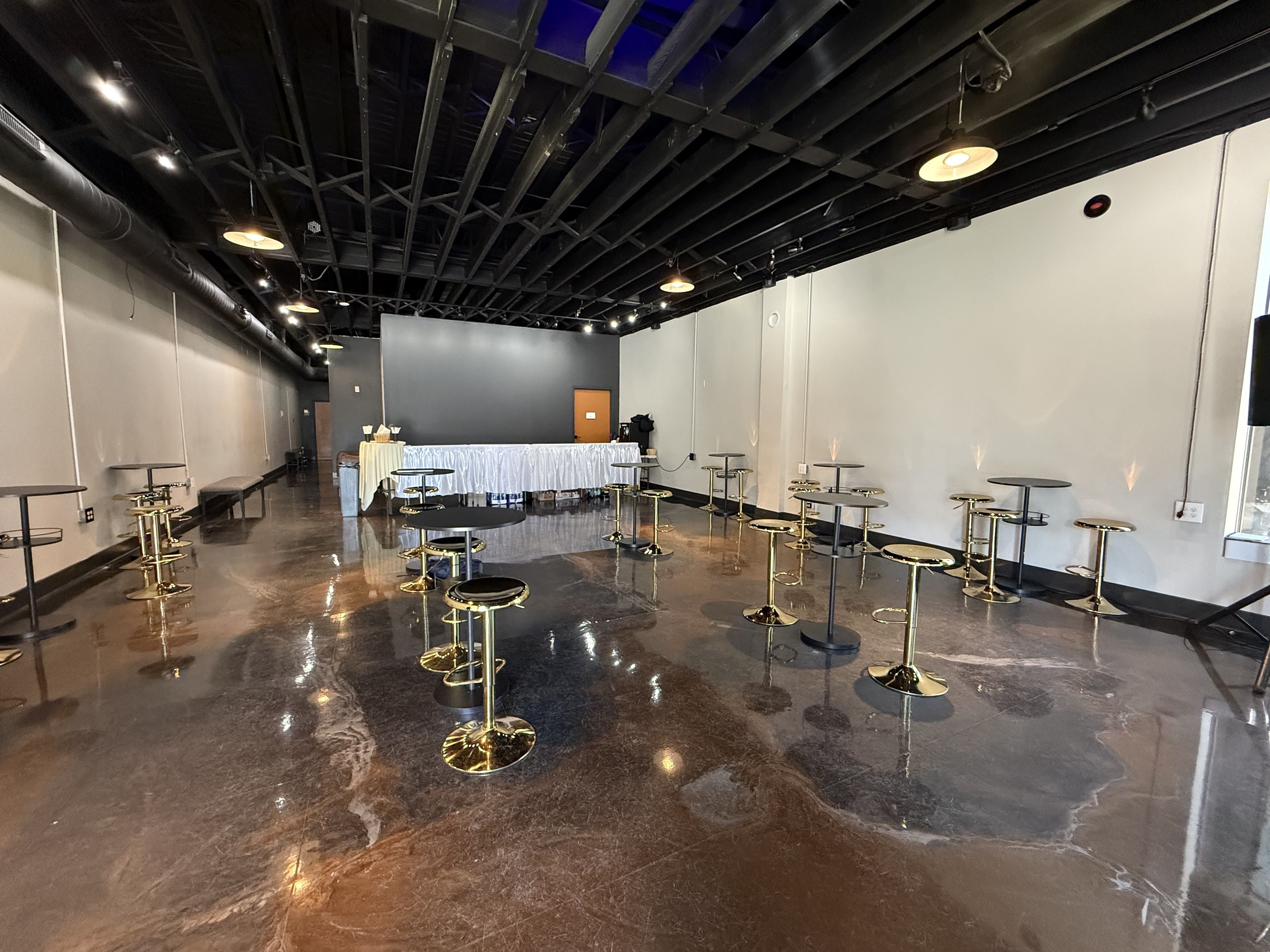 An industrial event space with a glossy concrete floor, black exposed ceiling, and warm pendant lights. Gold-based high tables are scattered around; at the far end a long table with a white skirt sits before a dark wall.
