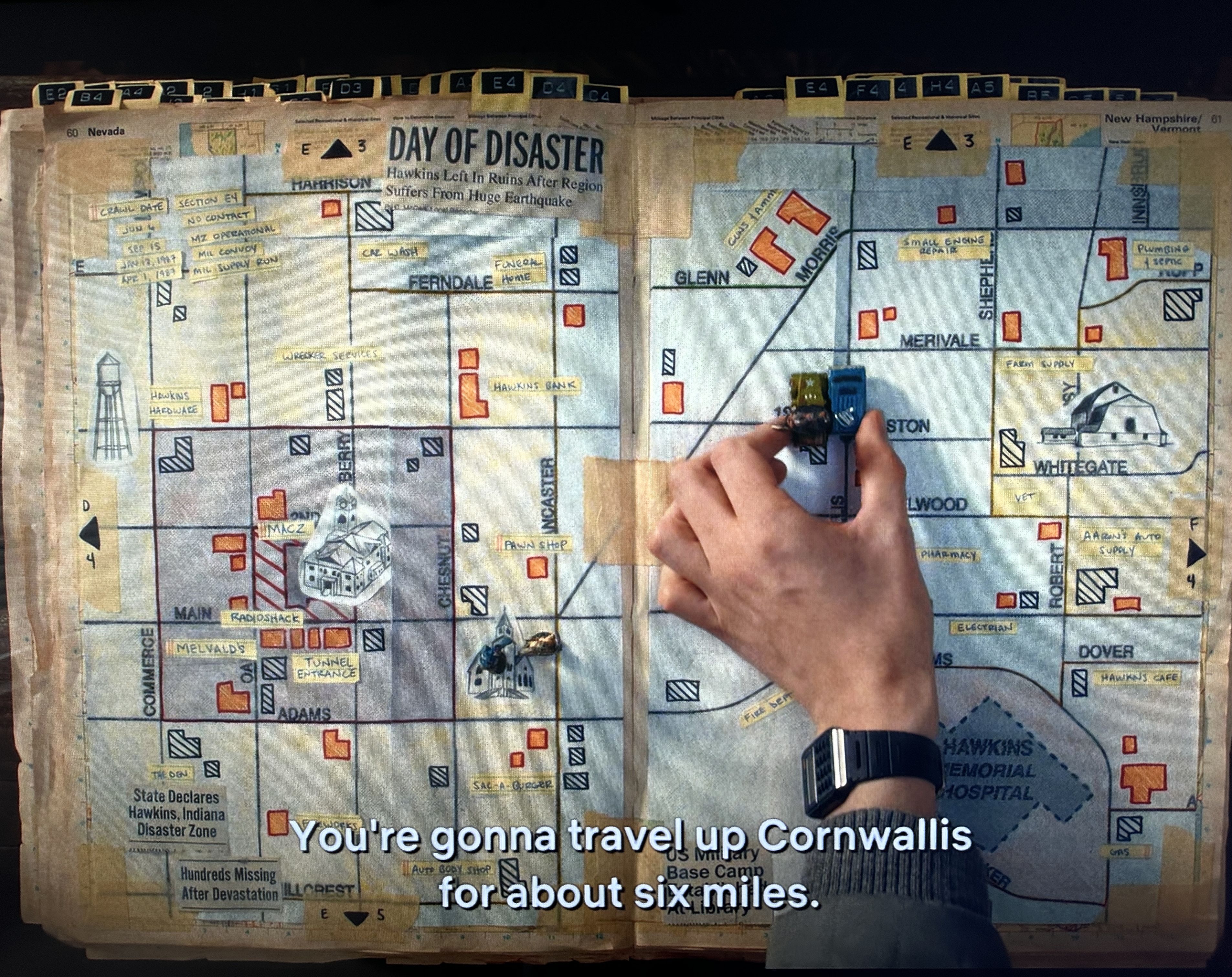 A hand placing blue and green toy trucks on an old, taped map labeled "DAY OF DISASTER" showing Hawkins, Indiana, with various locations like Hawkins Bank and Hawkins Hardware marked in red and yellow.