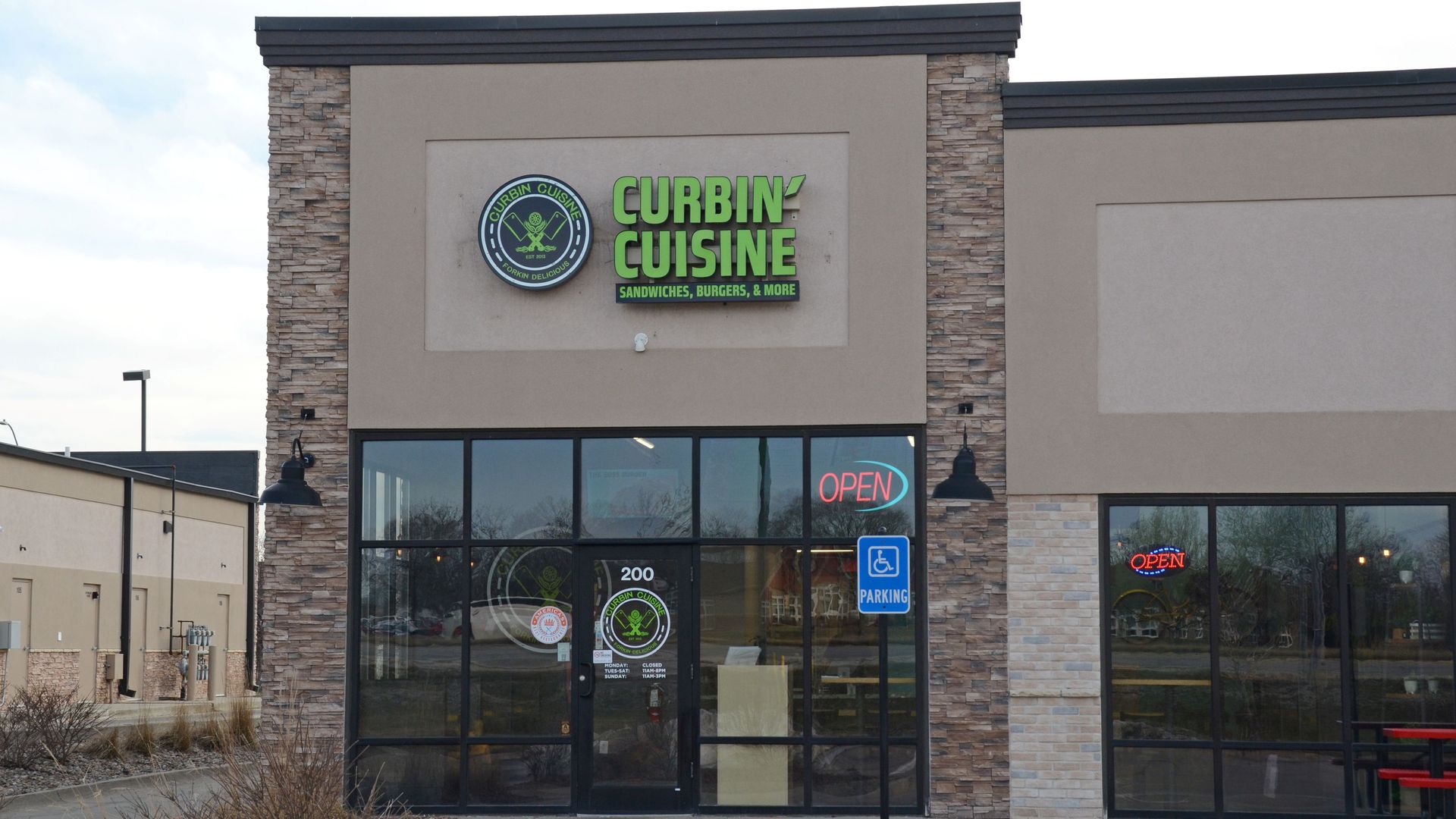 Exterior of Curbin' Cuisine restaurant with stone and beige walls, large glass windows, green signage, and red neon "OPEN" sign. Handicap parking sign in front.