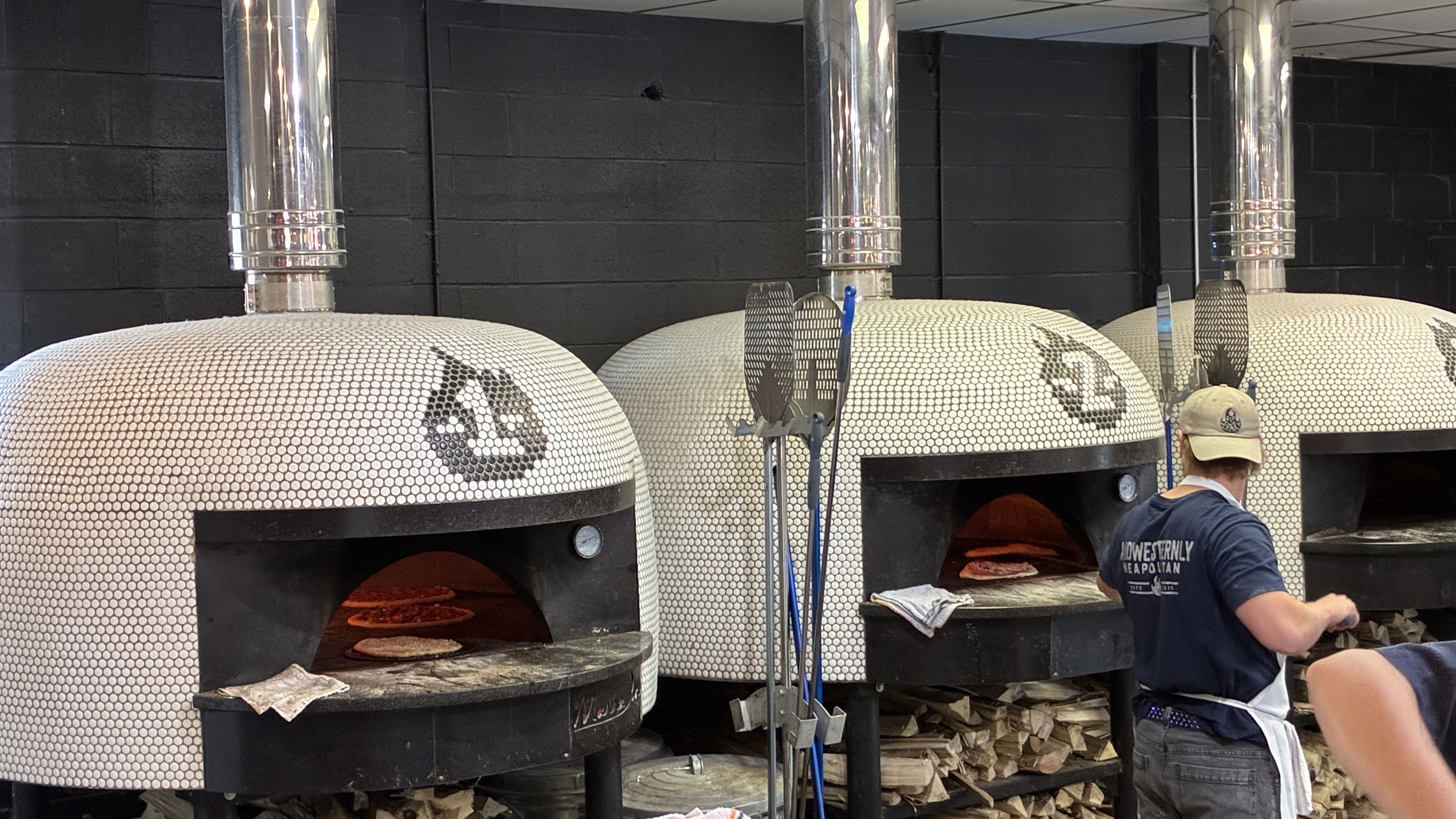 Three white mosaic-tiled wood-fired pizza ovens with tall shiny metal chimneys stand against a black brick wall; glowing pizzas bake inside. A worker in a cap and apron handles a pie.