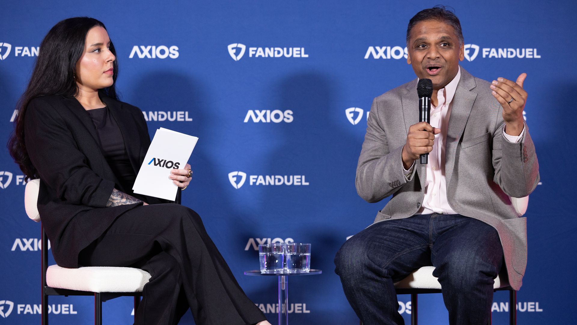 Two people seated on stage in front of a blue backdrop with Axios and FanDuel logos. The woman on the left holds an Axios folder, the man on the right speaks into a microphone.