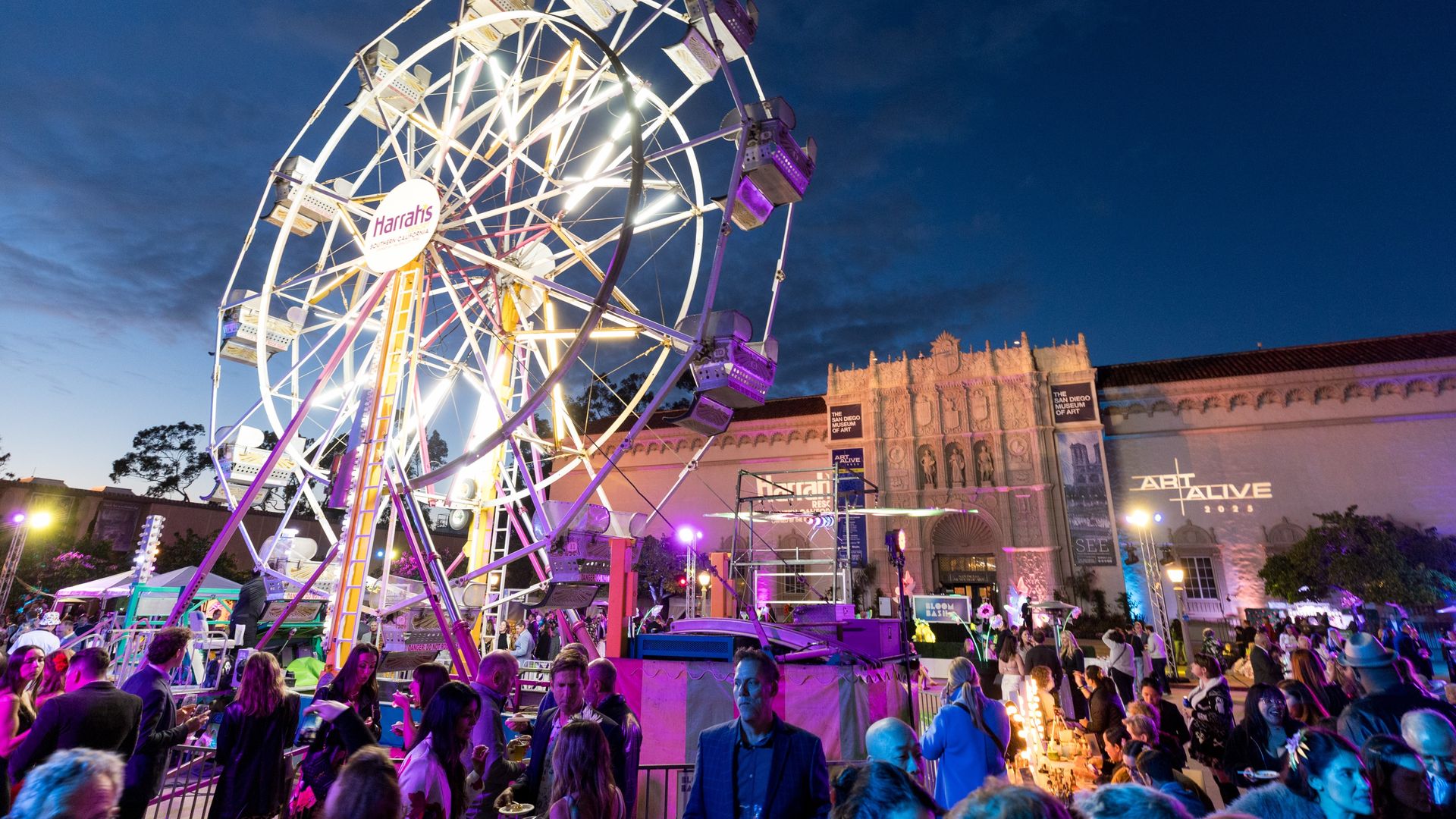 Nighttime festival scene with a large illuminated Ferris wheel labeled "Harrah's", white and purple frame, crowds at tables, and a lit historic building in the background.