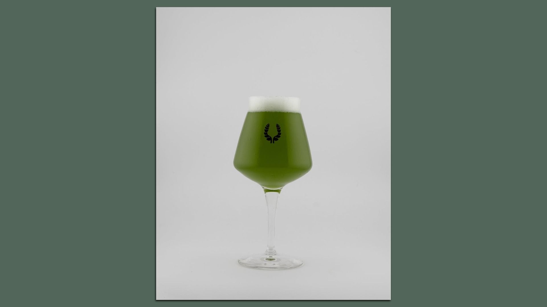 A green beer in a tall stemmed glass with a white foam head; a black laurel wreath logo is centered on the glass, set against a light gray backdrop.