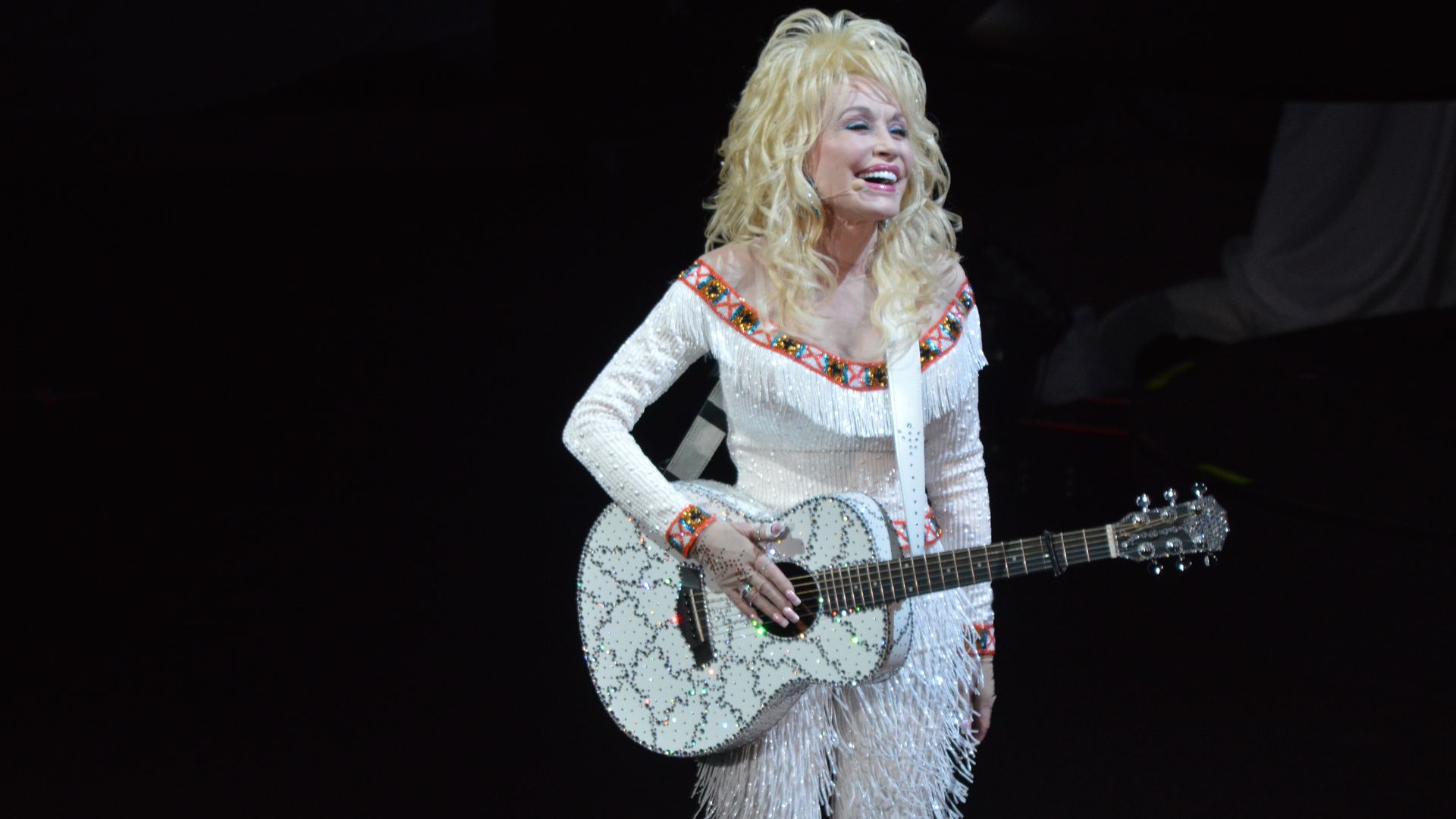 A female country singer with long blonde hair in a sparkling white fringed outfit, playing a rhinestone-studded white guitar on a dark stage, smiling.