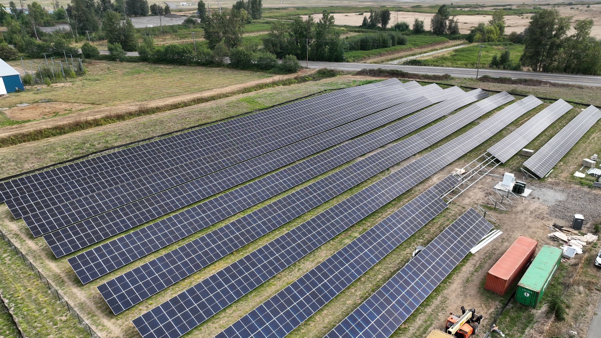 Aerial view of a large solar panel farm with multiple rows of dark blue panels on green grass, surrounded by trees and fields, with some shipping containers and equipment nearby.