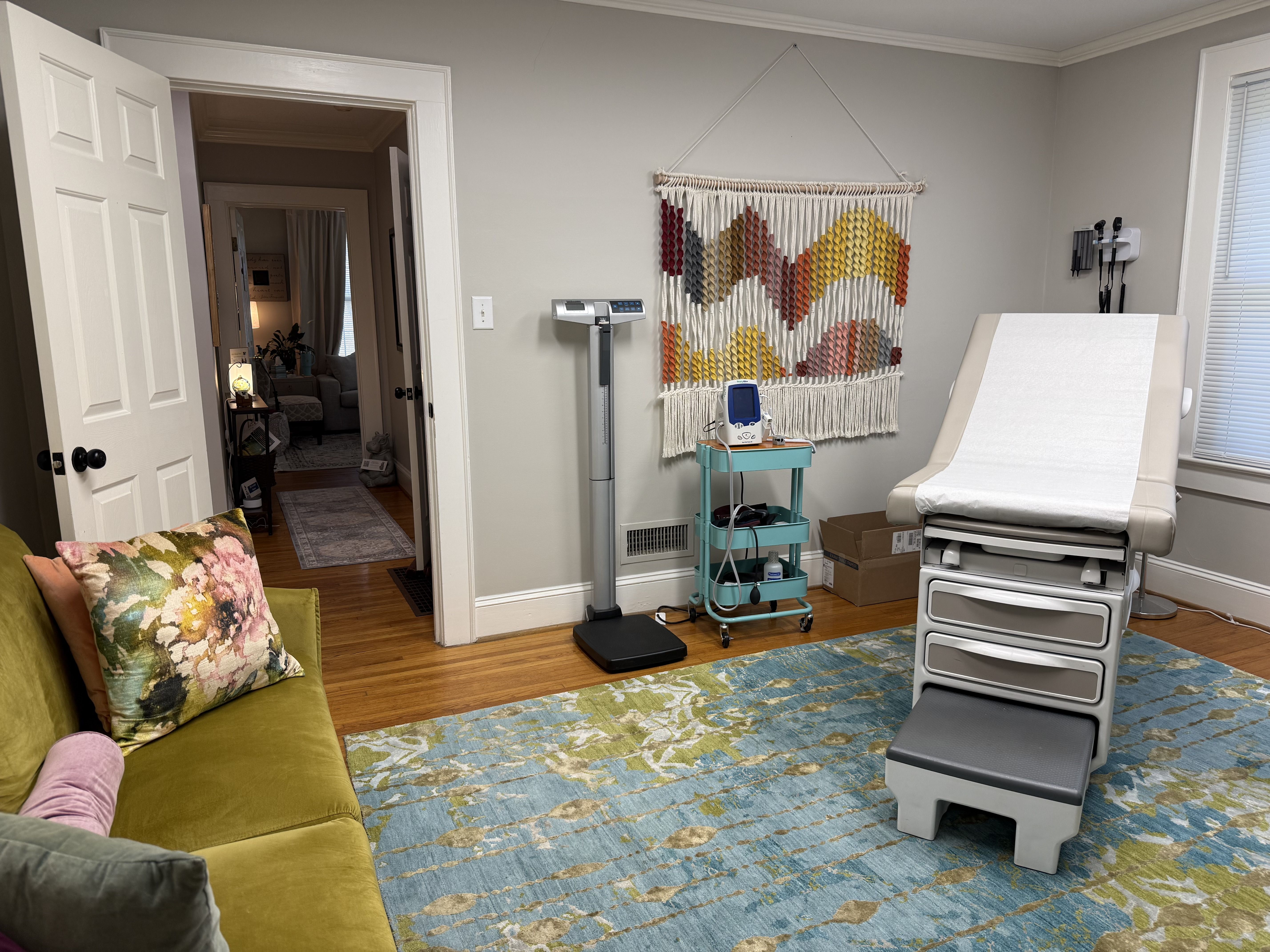Medical exam room with a white patient bed on the right, a teal cart and tall scale, wall equipment, and a colorful geometric wall tapestry; doorway reveals a living room beyond.