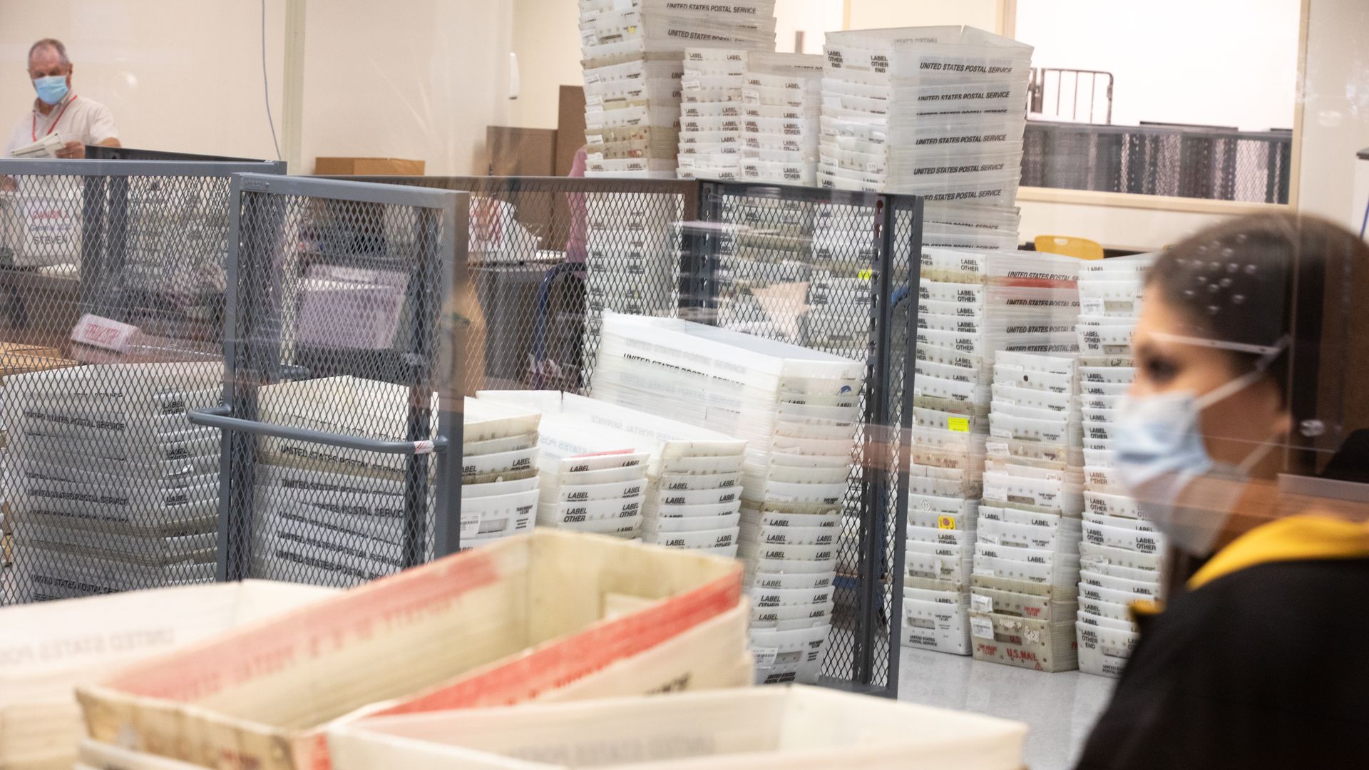 An election worker stands in a room with dozens of containers of uncounted ballots. 