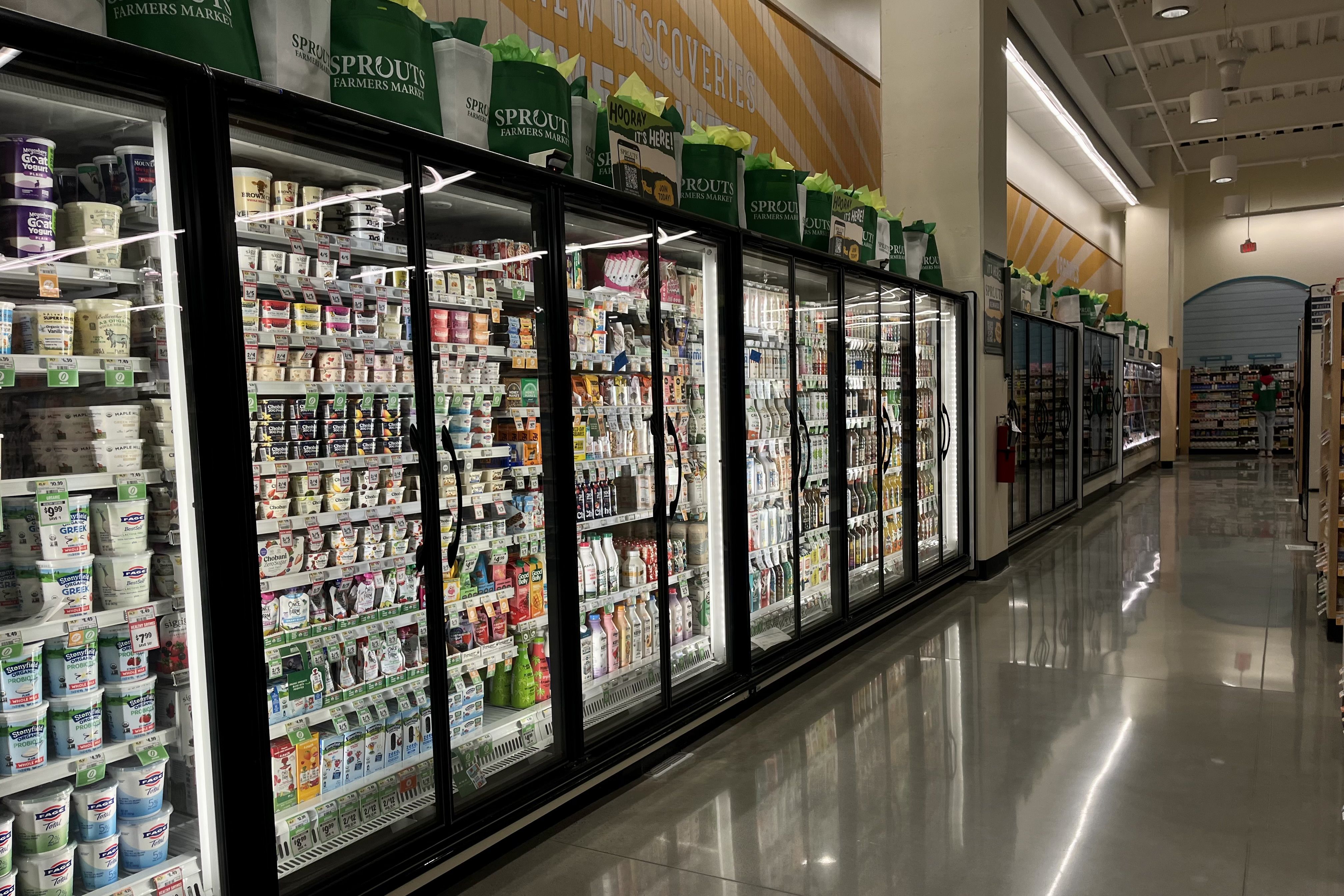 A grocery store aisle with glass-door refrigerators filled with various dairy products like yogurt and milk. Green Sprouts Farmers Market bags line the top shelf under yellow wall signage.