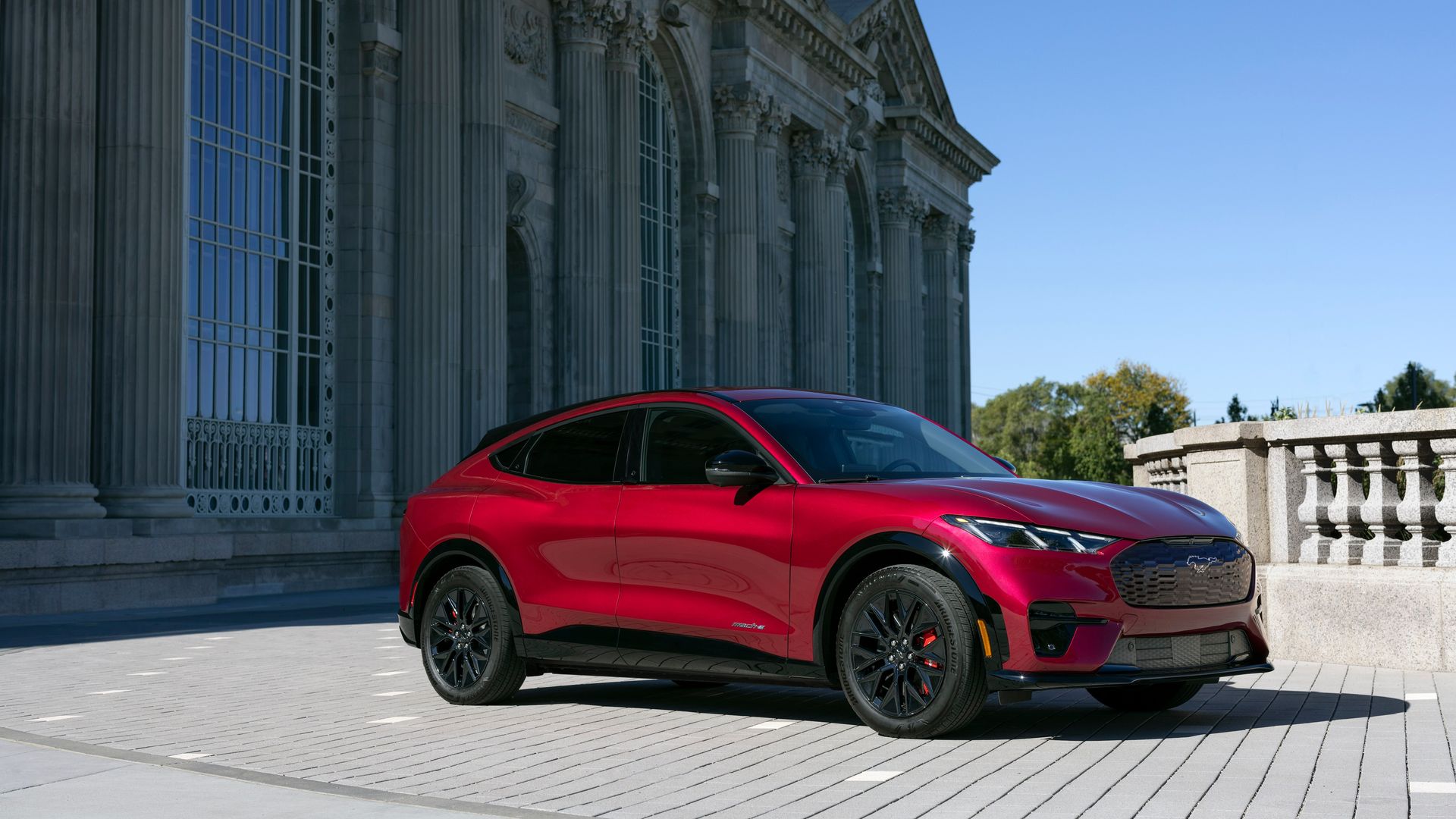 Shiny red Ford Mustang Mach-E electric SUV parked on grey brick pavement in front of a large classical stone building with tall columns and arched windows under clear blue sky.