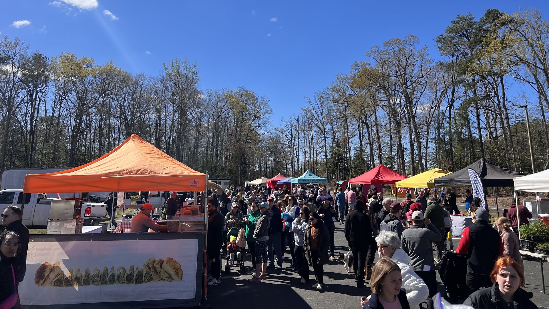 Crowds of people walking through a market with colorful tents up. One is orange and on the left side with a pic of empanadas put up 