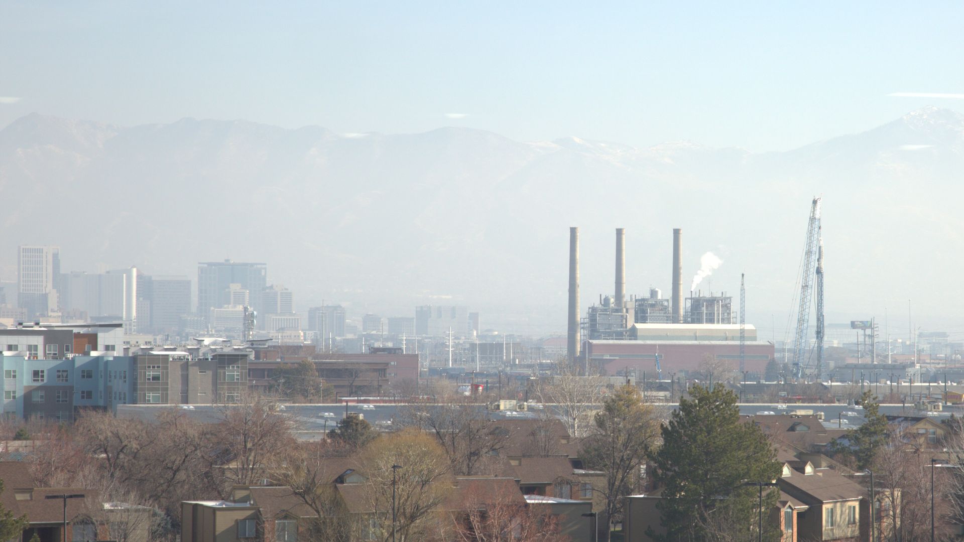 Cityscape with buildings, industrial smokestacks emitting white smoke, and taller city skyscrapers in haze. Mountains are faintly visible in background beneath clear blue sky.