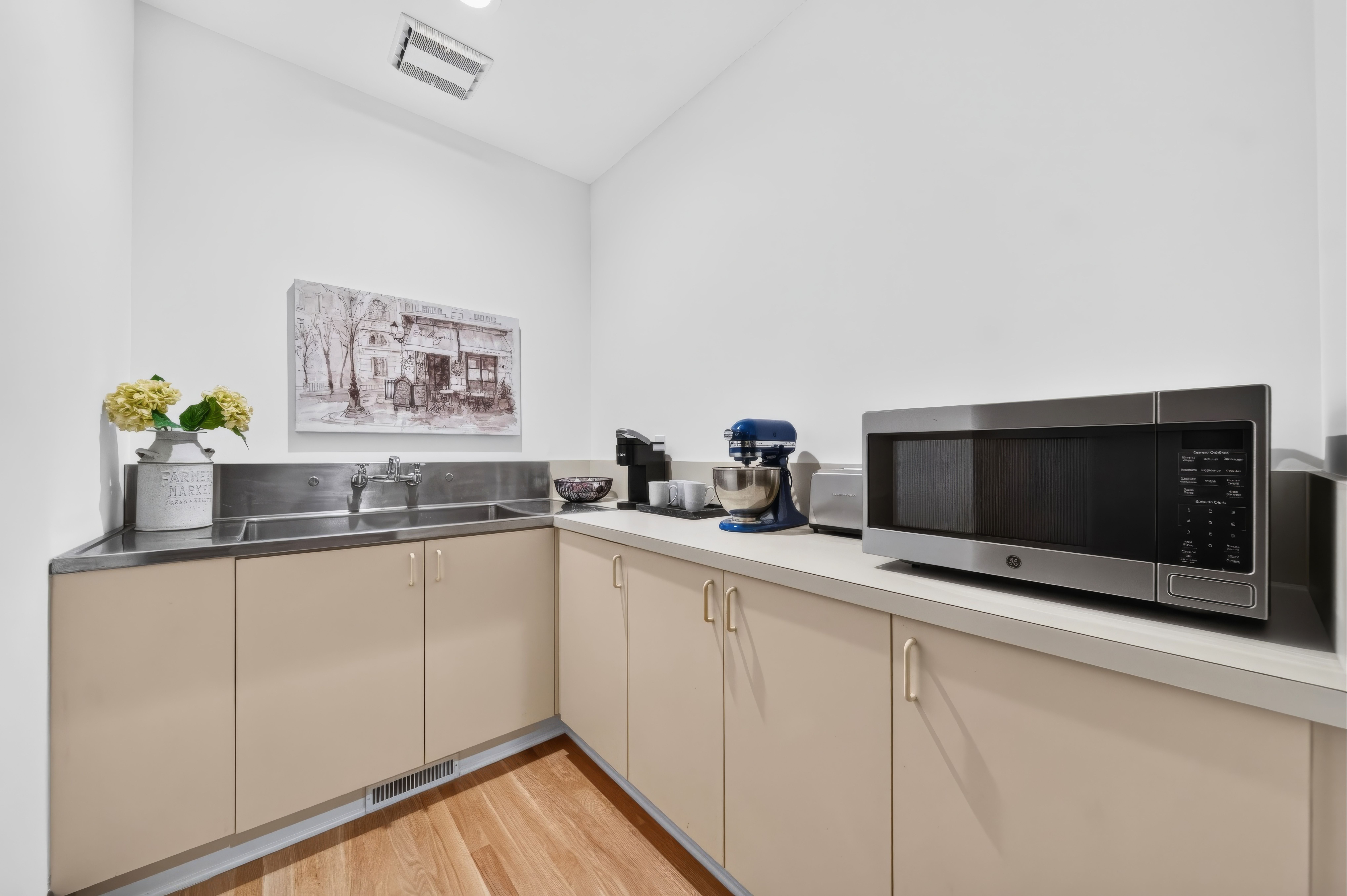 Bright kitchen with beige cabinets and stainless counters. A blue stand mixer sits near a microwave; a black-and-white cityscape painting hangs above the counter, with yellow flowers on left.