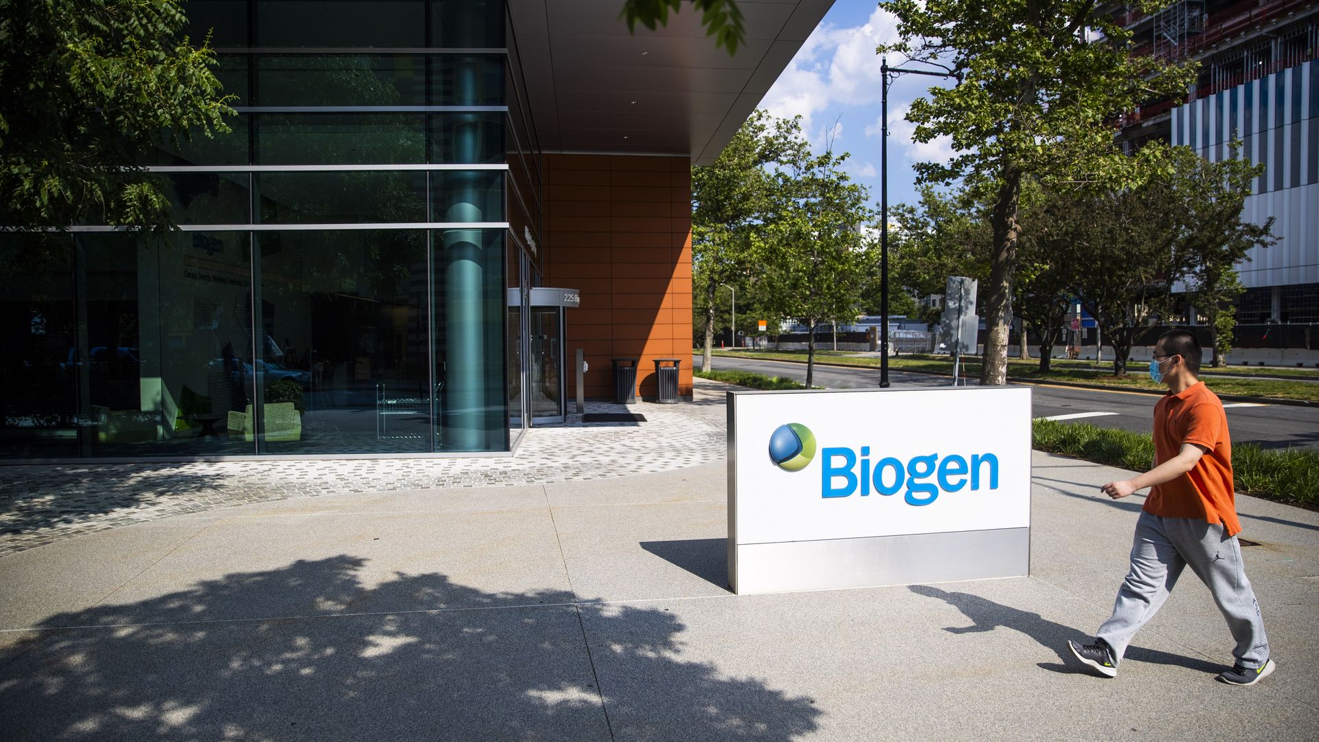 A man walks in front of a blue and white Biogen sign and the Biogen headquarters building.