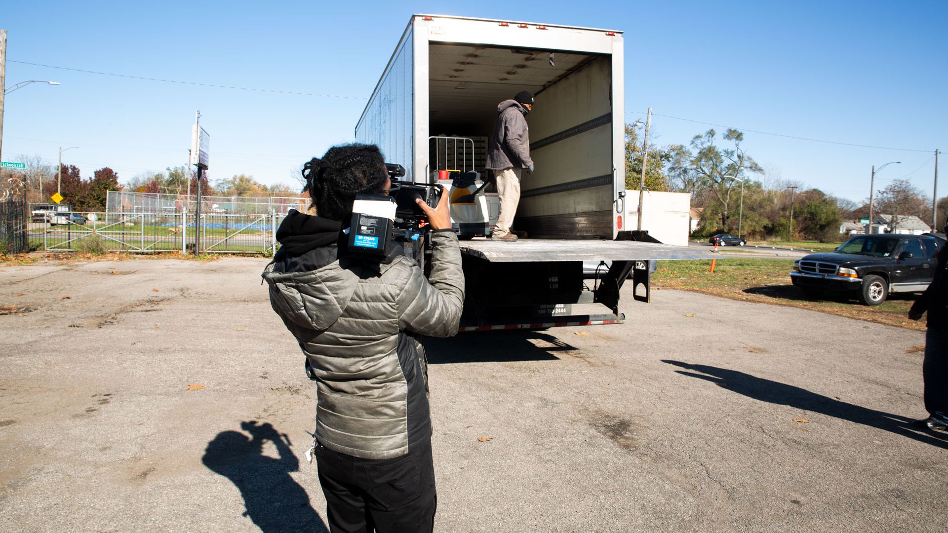 A cameraperson in a gray puffer jacket films a man in a gray jacket and beanie stepping into the back of a large white box truck with its rear doors open.