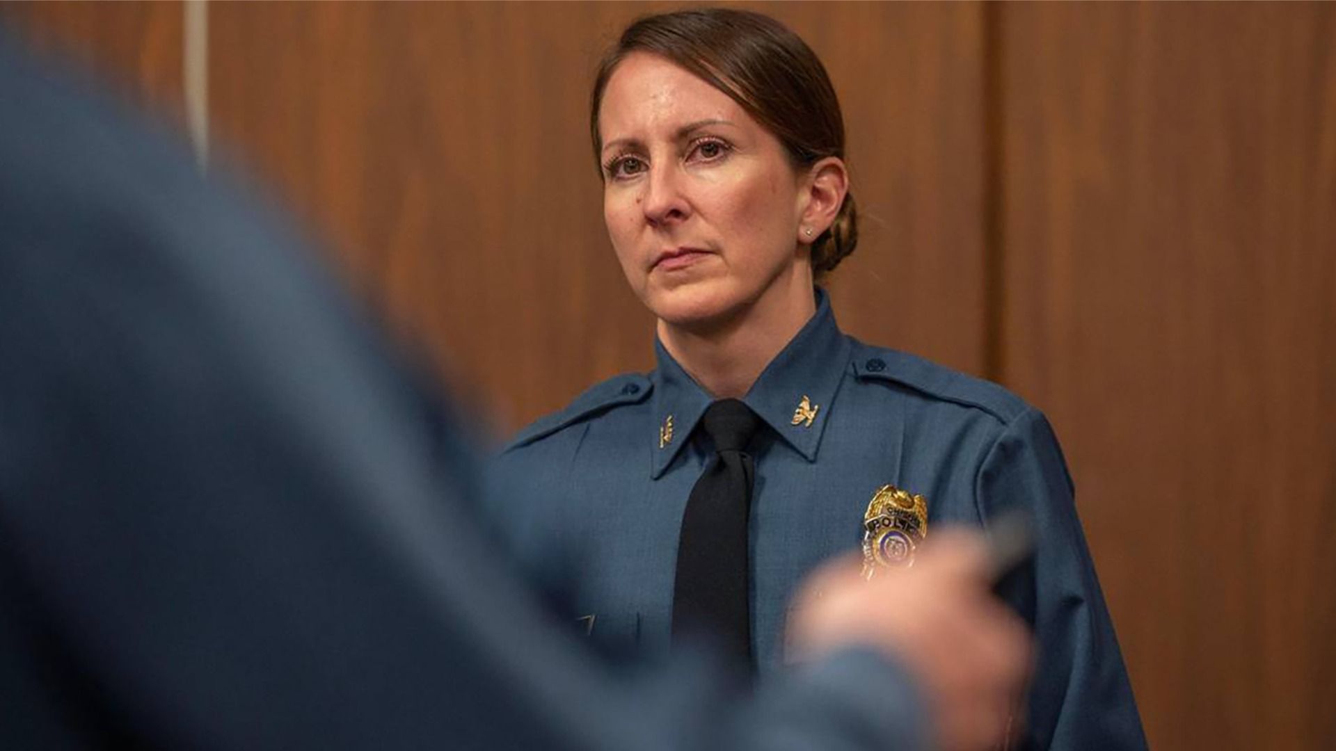 Kansas City Police Chief Stacey Graves standing in a blue shirt police uniform with a badge and a black tie, with a concerned facial expression in front of a wood panel wall.