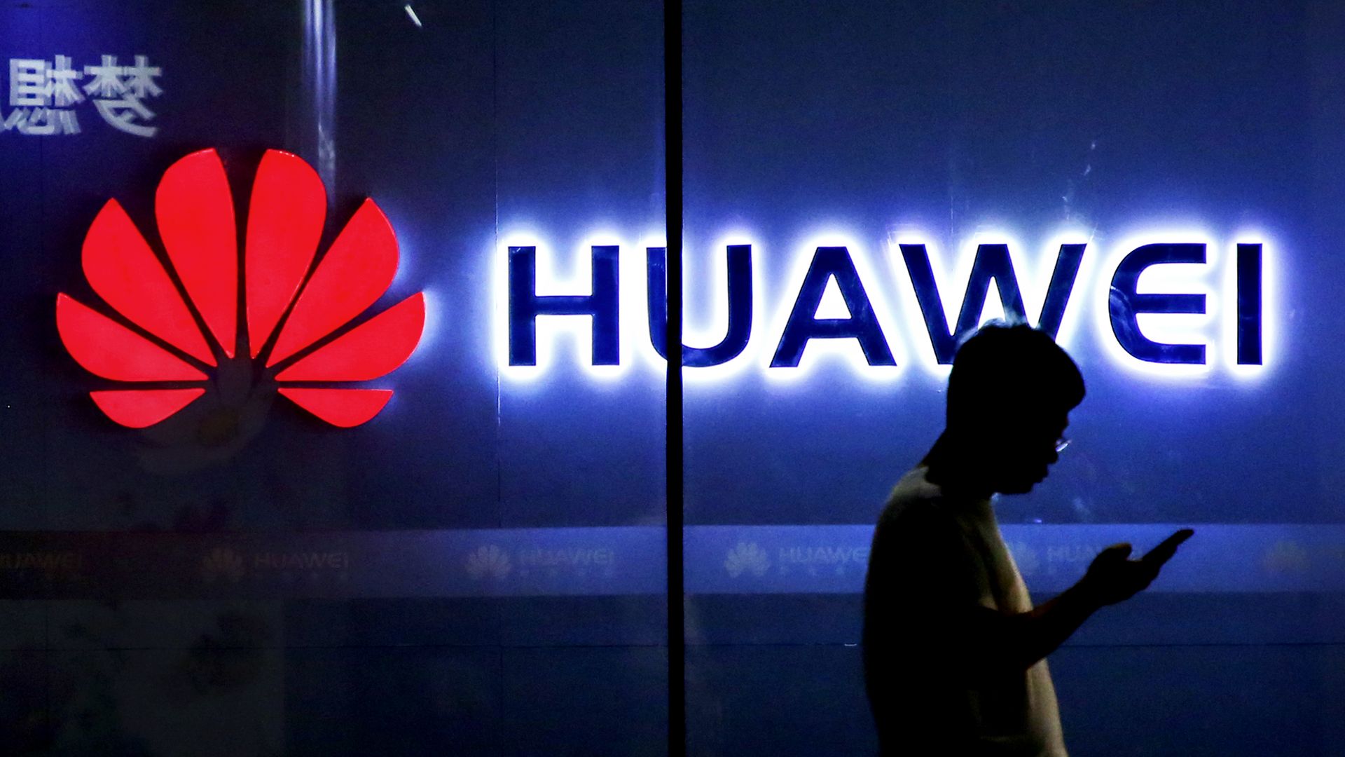 Photo of a man with a smartphone silhouetted against a glowing Huawei sign