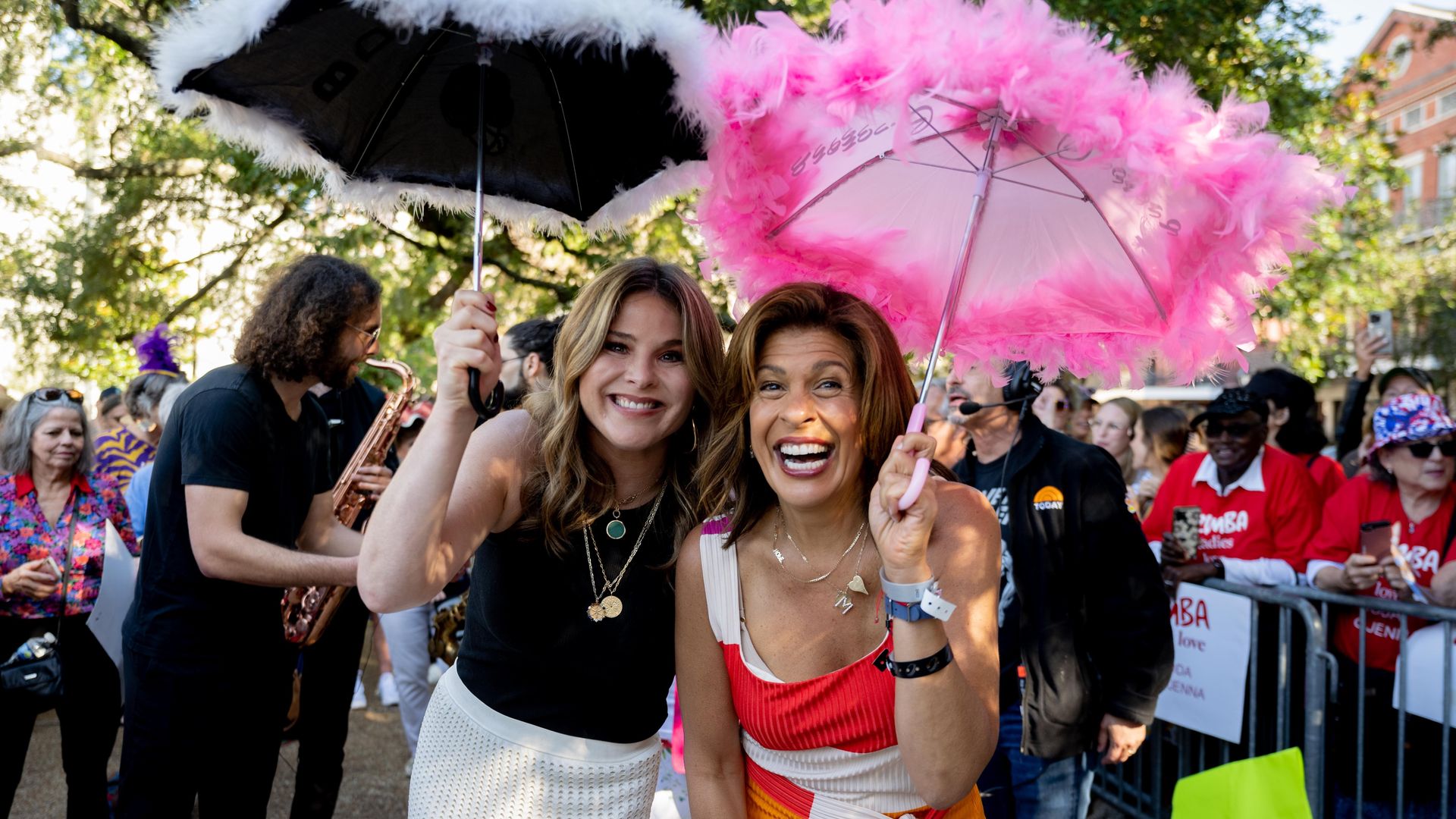 Photo shows Jenna Bush Hager with a black parasol trimmed in white feathers. Hoda Kotb is smiling with a pink parasol with pink feathers. A saxophone musician plays behind them.
