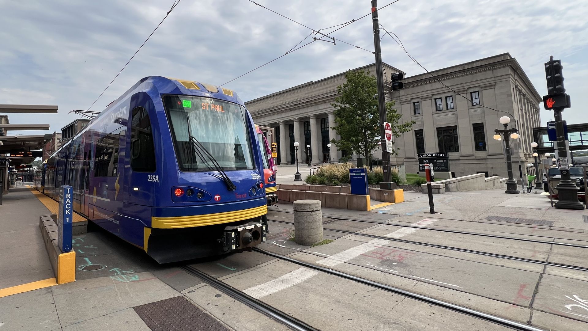 A light rail train car with blue trim rolls into an outdoor transit station near a large neoclassical stone building with a large columned entryway