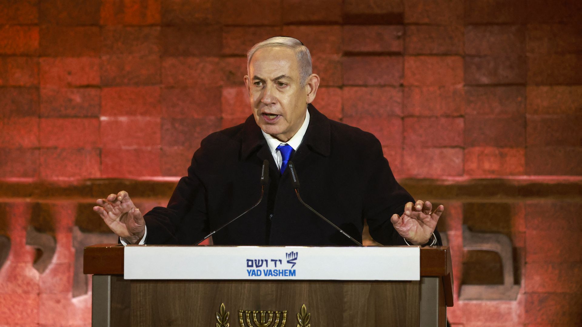Benjamin Netanyahu wearing a black coat and speaking in front of a brick wall at a wood podium.