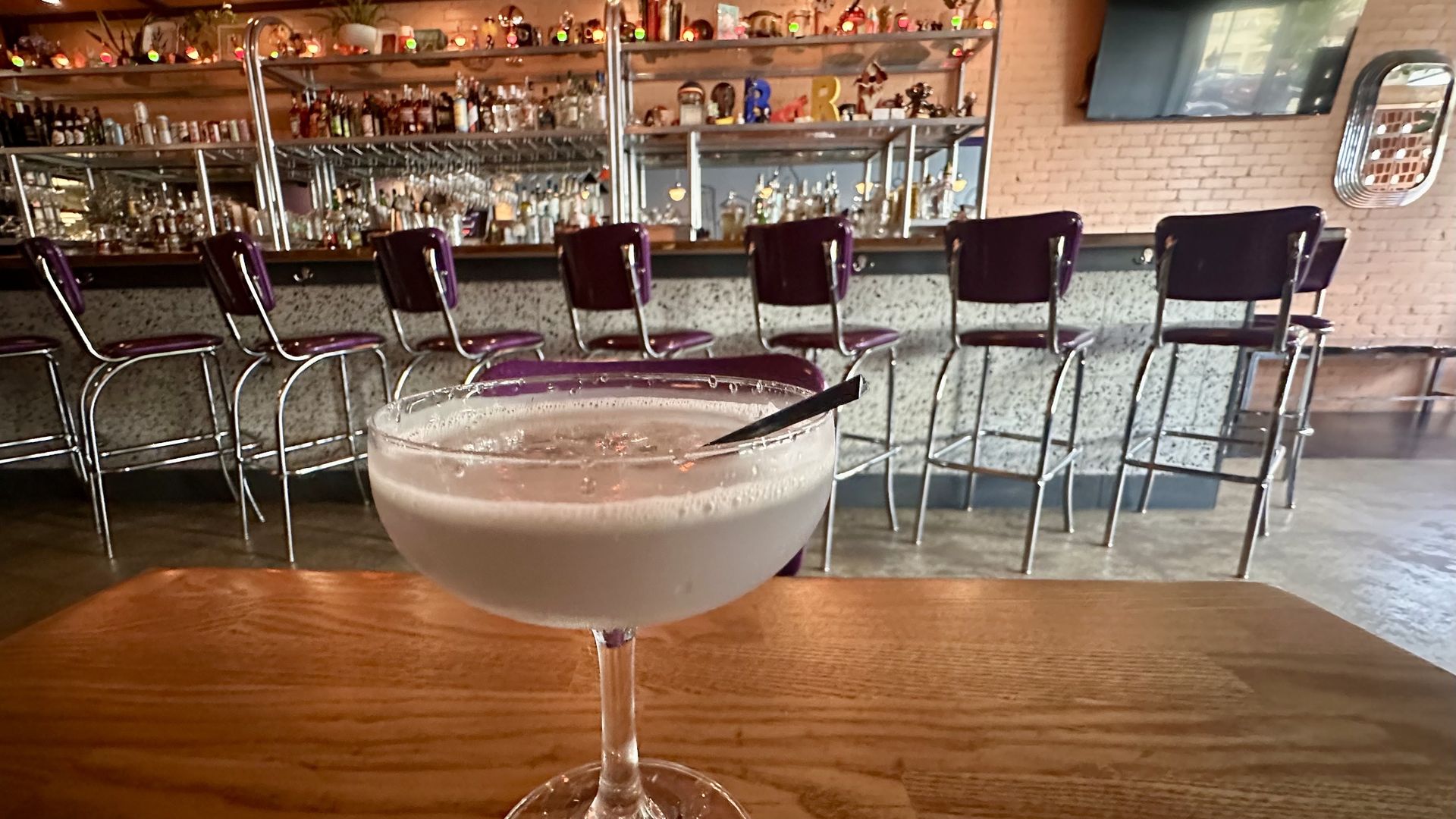 A photo of a purple-ish cocktail in a coupe glass on a table in front of a bar with retro tall diner seats
