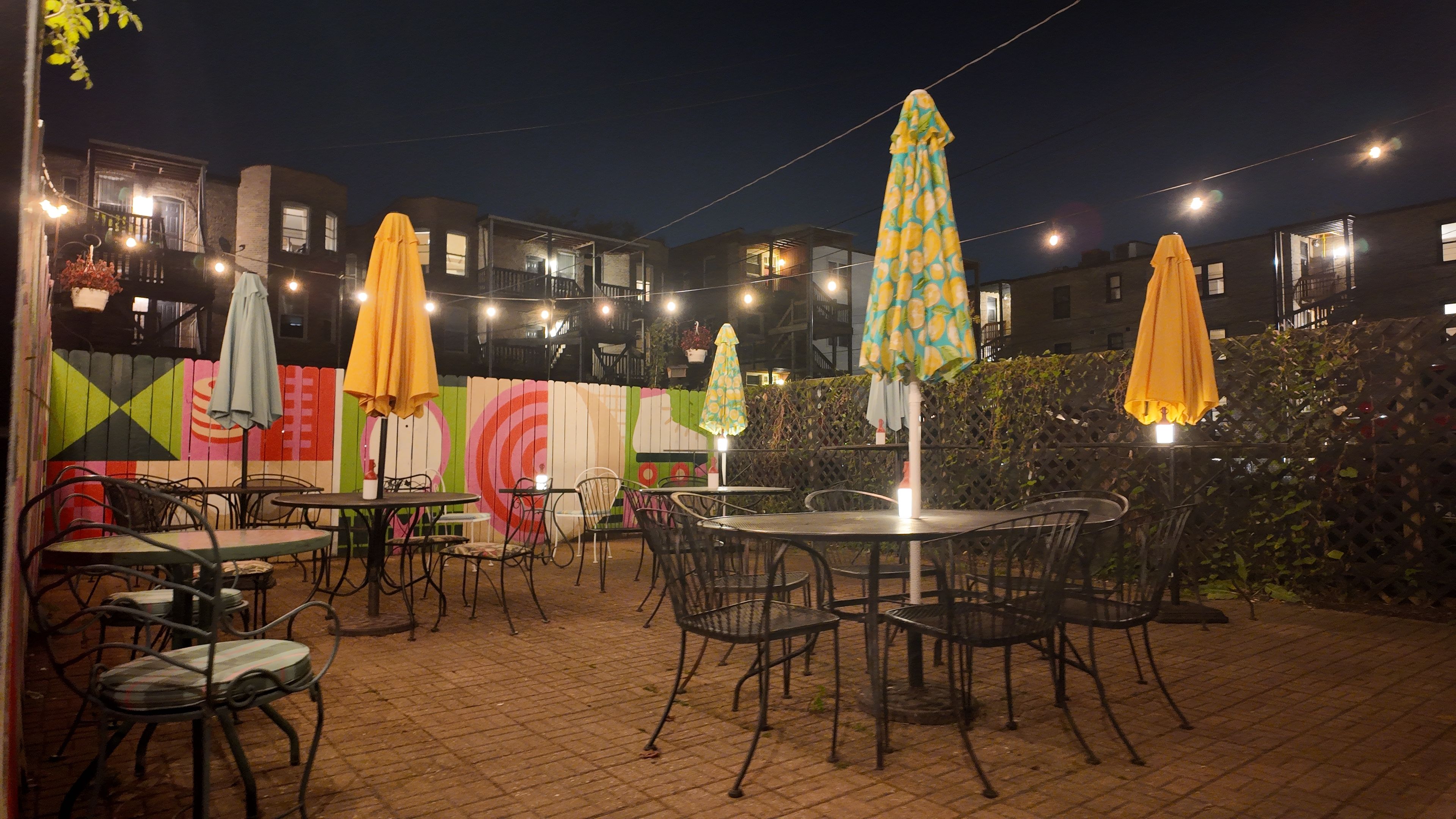 Outdoor patio at night with metal tables and chairs, colorful closed umbrellas, string lights overhead, and a vibrant mural on the wooden fence behind surrounded by apartment buildings.