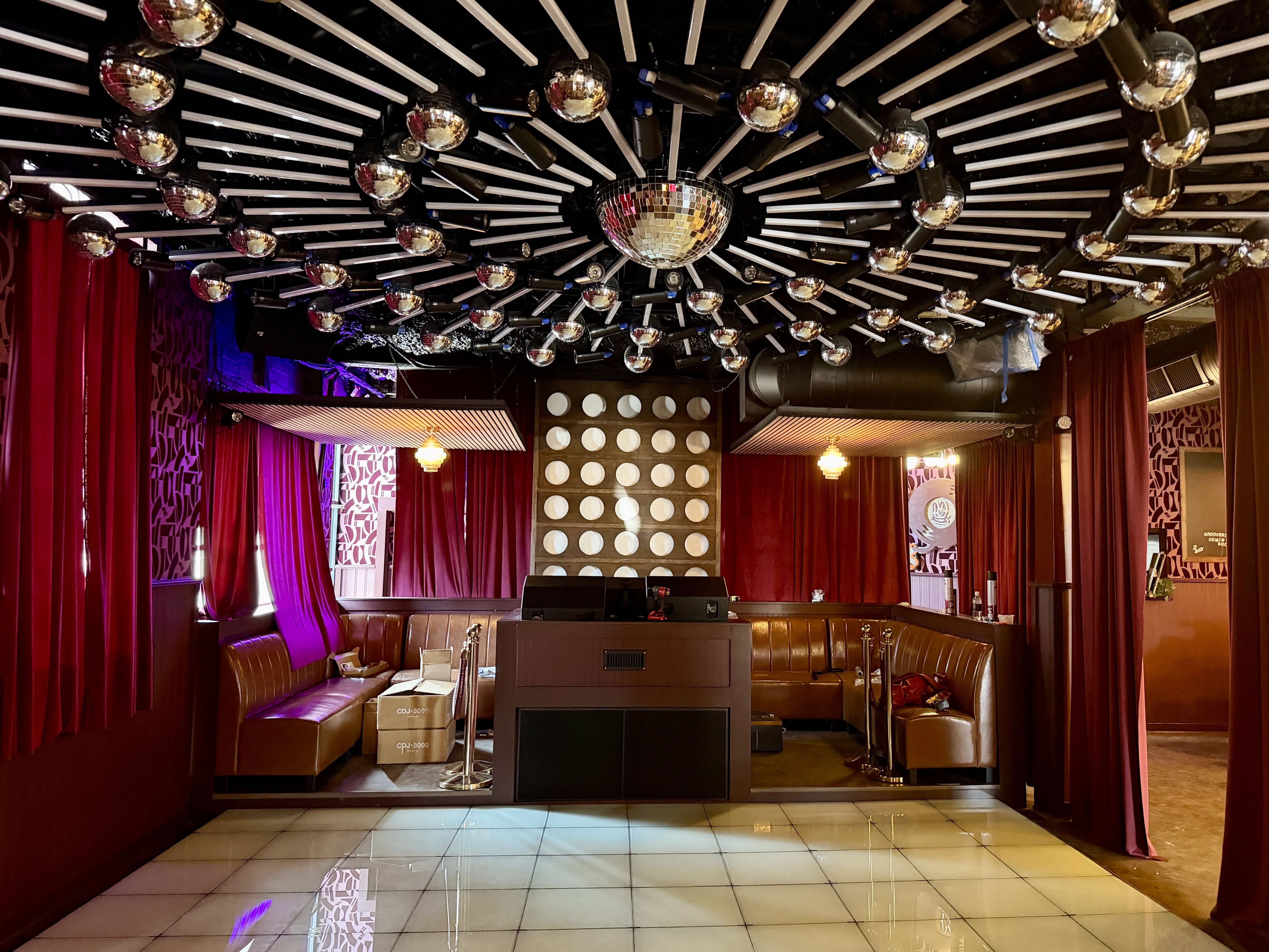 Interior of a nightclub or lounge with red curtains, brown leather seating, a mirrored disco ball ceiling installation, and a central DJ booth on a tiled floor.