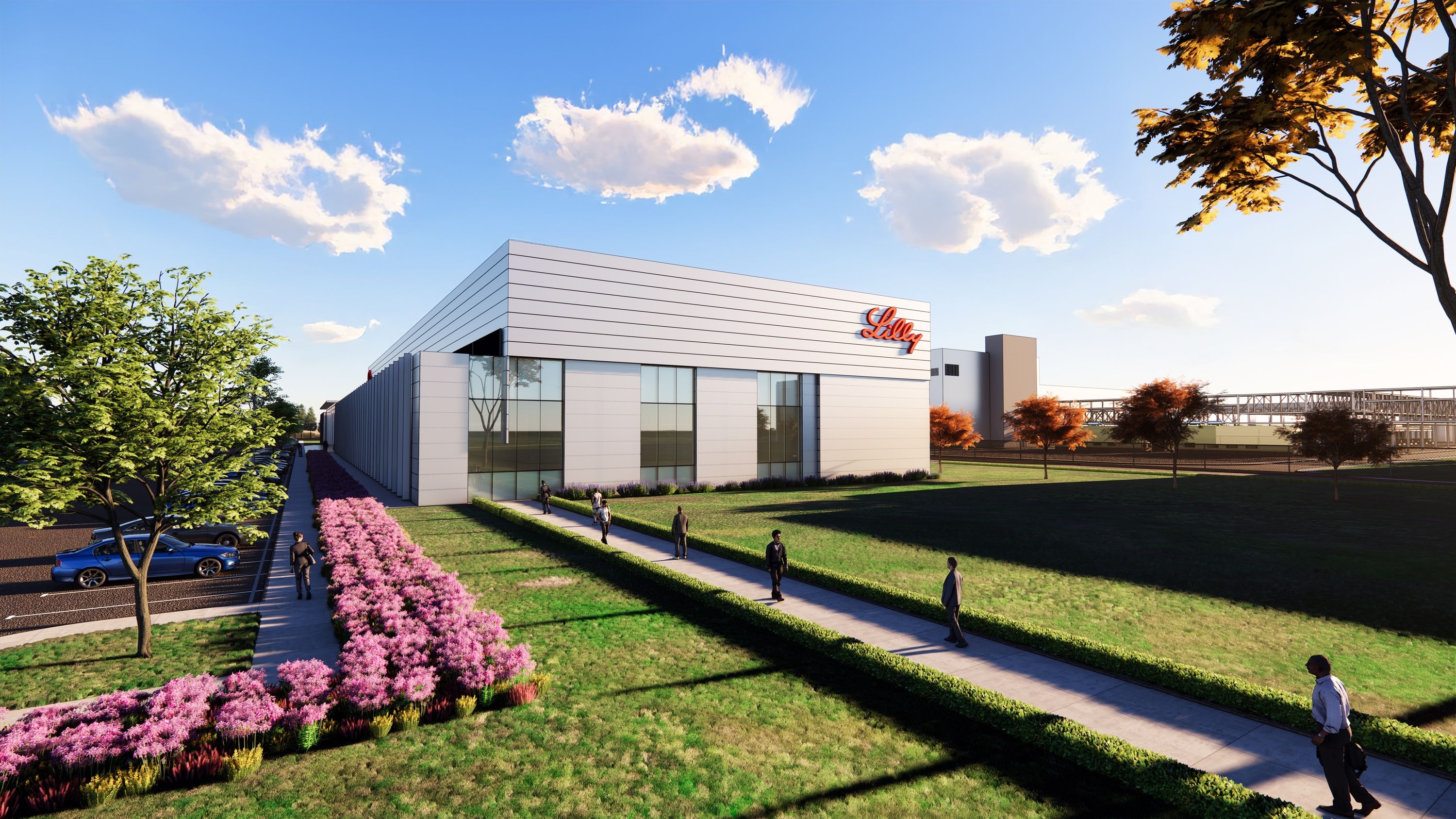 Modern white building with the red Lilly logo on a sunny day, surrounded by green lawn, pink flowers, trees with autumn leaves, and people walking on concrete paths.