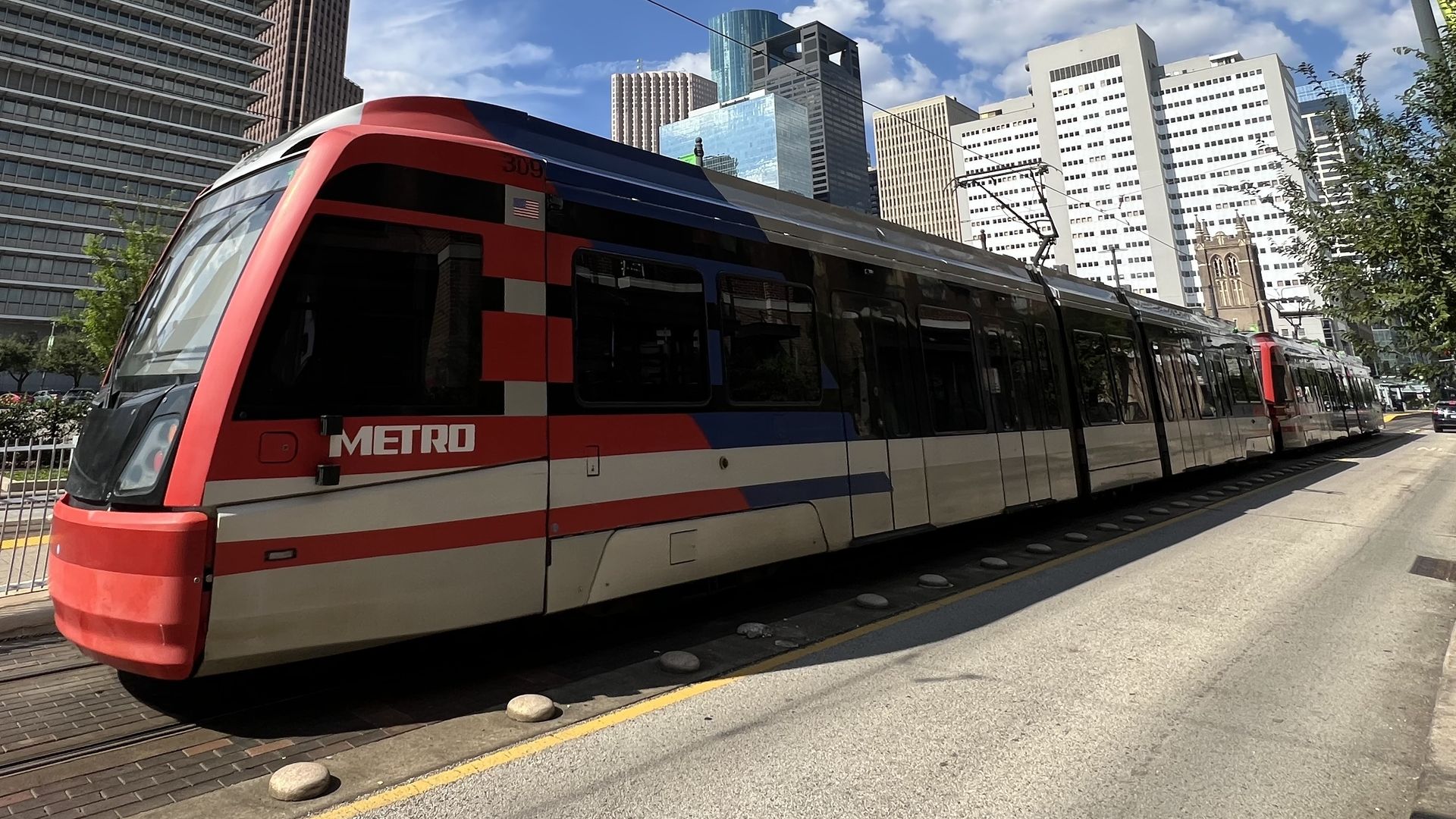 Red, white, and blue Metro light rail train on tracks in a city with tall skyscrapers under a partly cloudy blue sky.