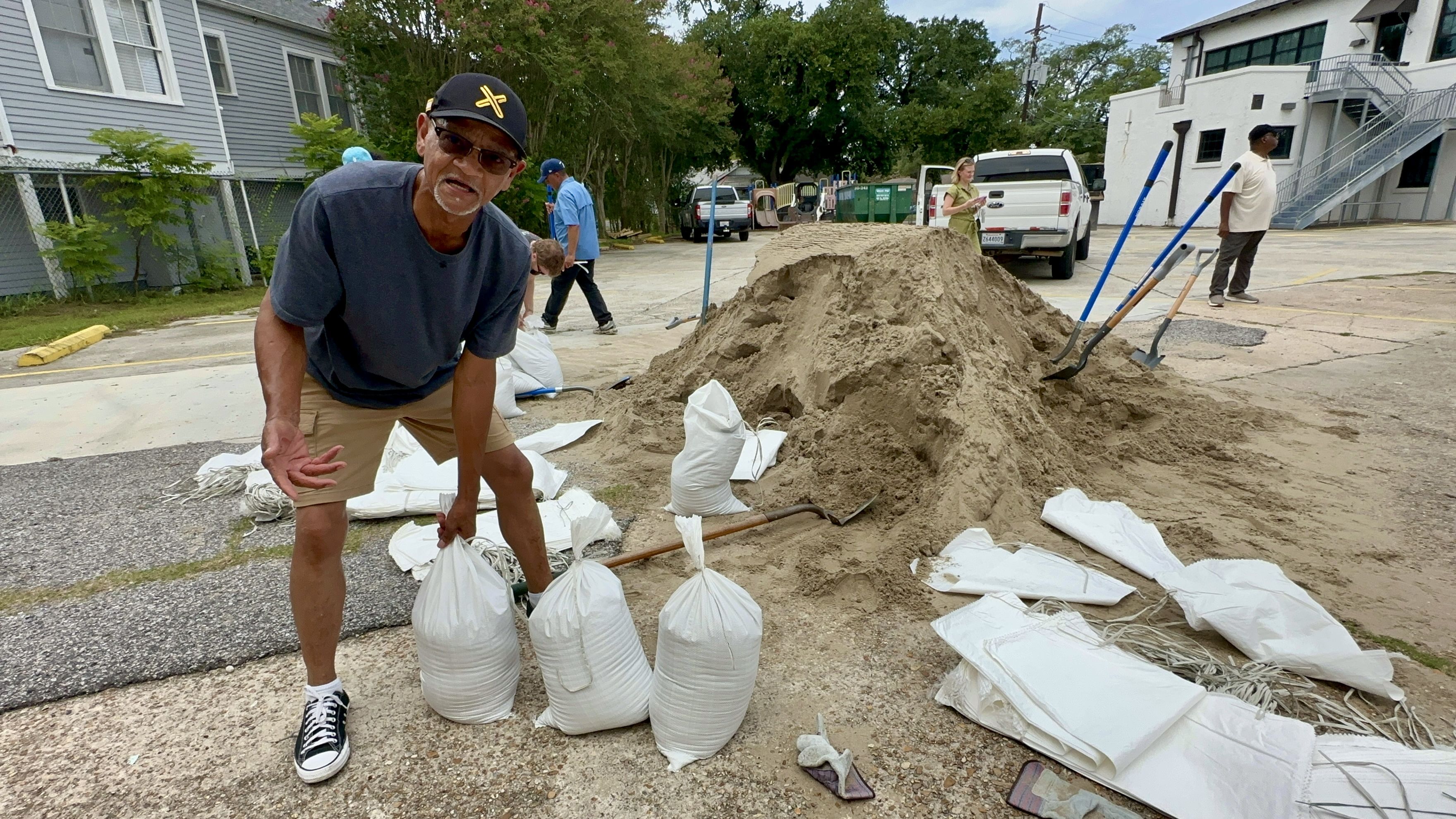 Man wearing black cap and glasses filling white sandbags from a large sand pile in an outdoor area with shovels and other people in the background.