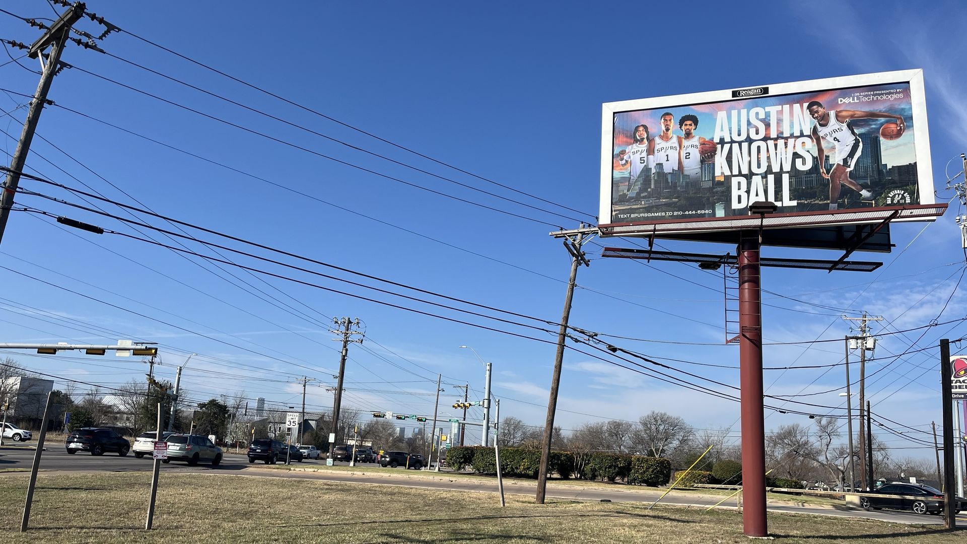 Billboard on a pole near a road with cars and traffic lights, showing four basketball players in Spurs jerseys with text "Austin Knows Ball" against a blue sky background.