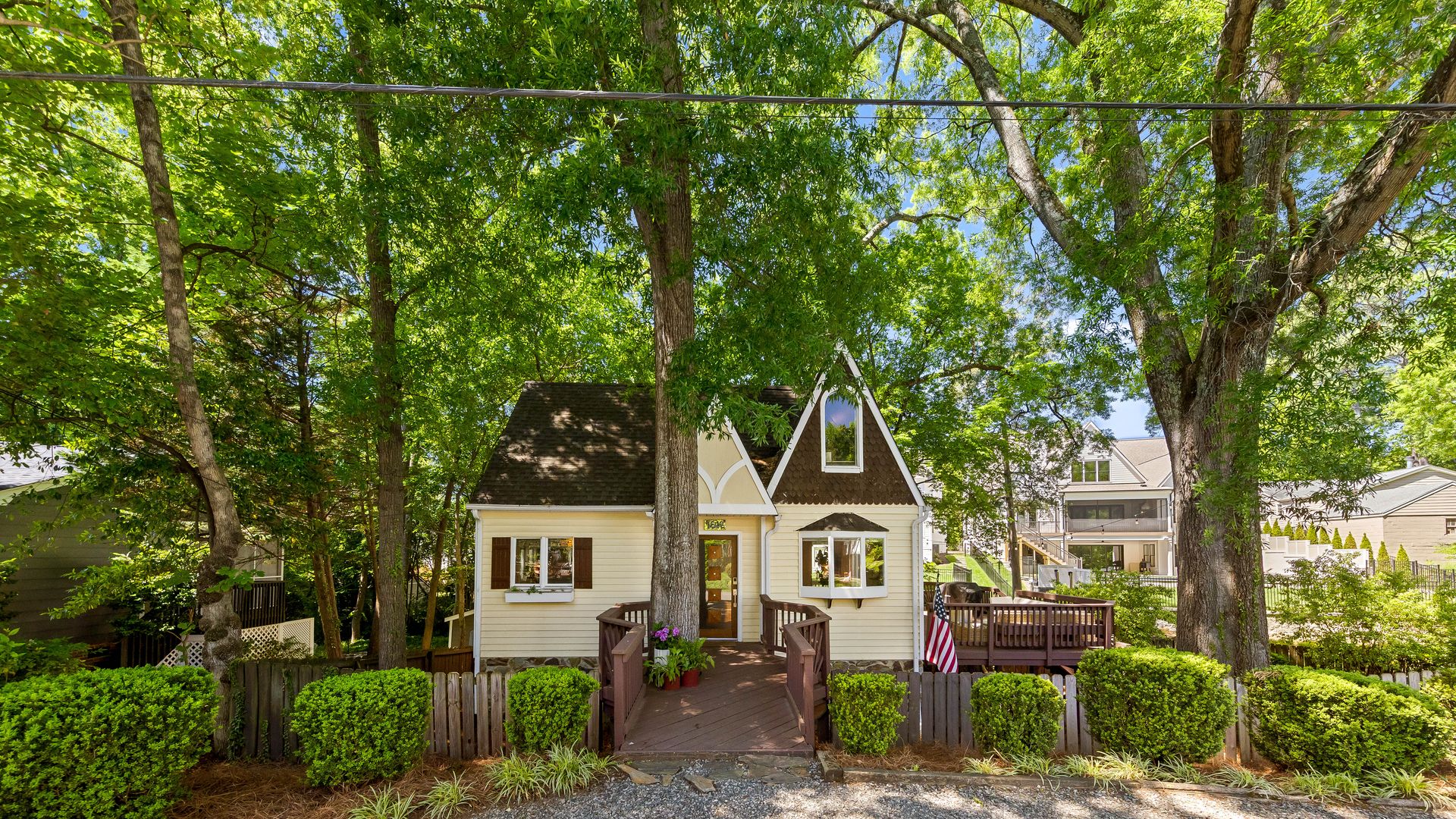 house for sale. Small cream house with a brown roof and triangular gables sits behind a wooden deck and fence, under a canopy of tall green trees; a gravel driveway and an American flag are visible to the right.
